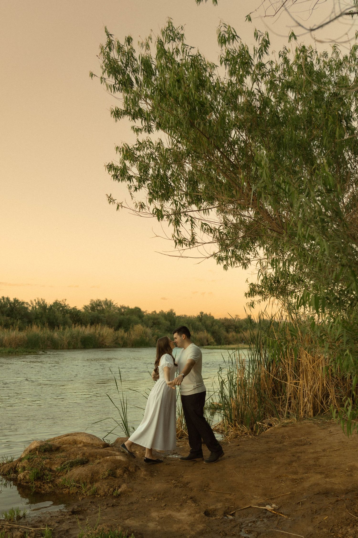 Couple embracing by a river at sunset, surrounded by lush green trees and reeds, woman in white dress.