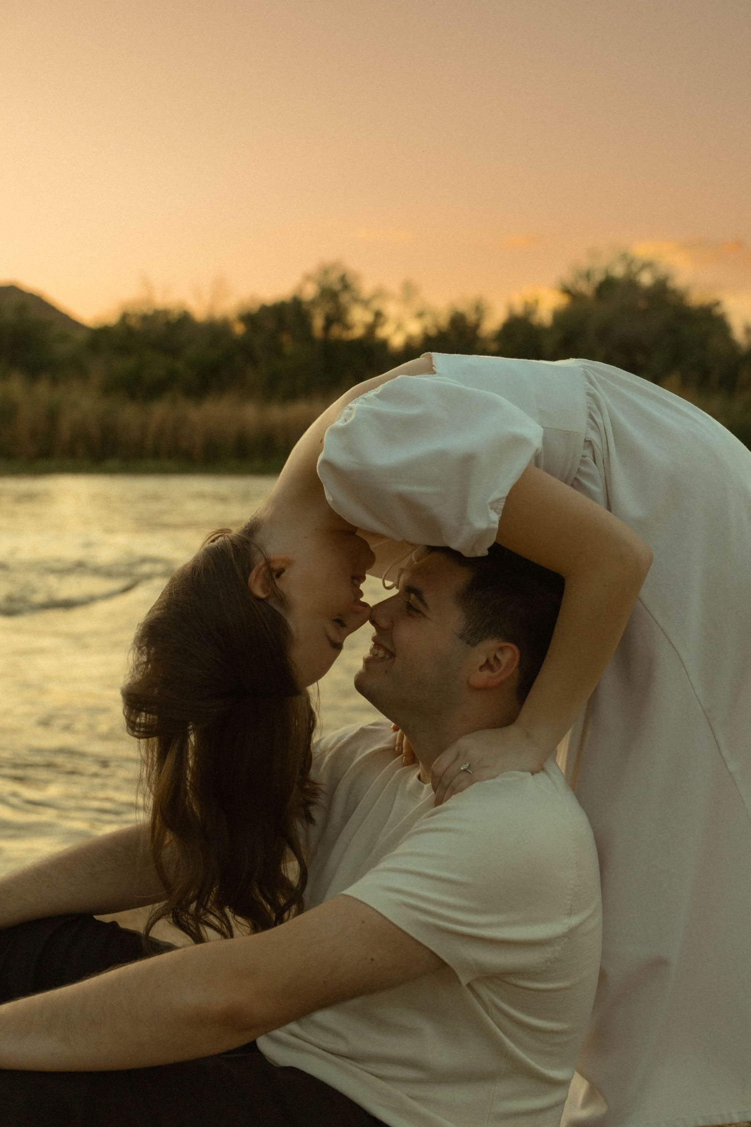 Romantic couple leaning in for a kiss at sunset by the river, woman in white dress arching back dramatically.