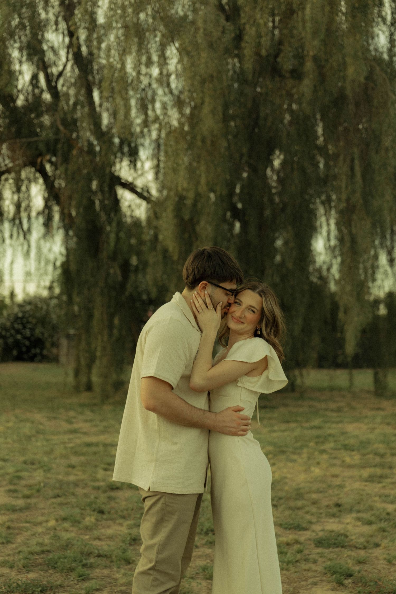 Happy couple hugging and smiling under a weeping willow tree in a green park.