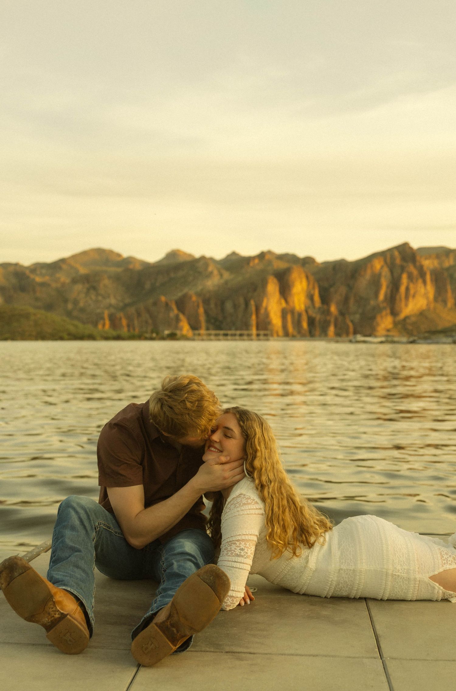 Intimate moment between a couple on a dock at sunset with golden cliffs and water in the background.