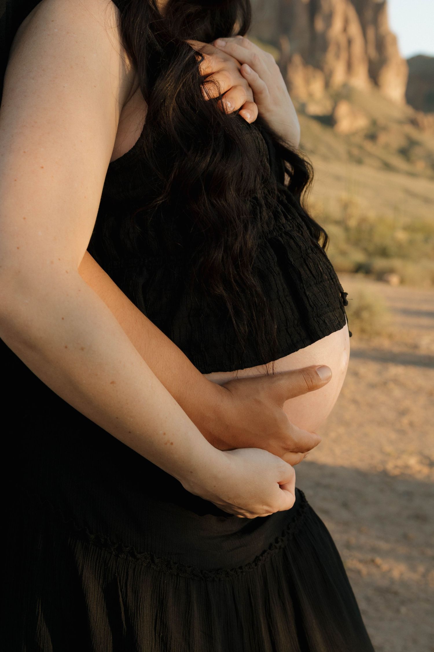 Close-up photo of a pregnant woman's belly in a black dress against a desert landscape at sunset.