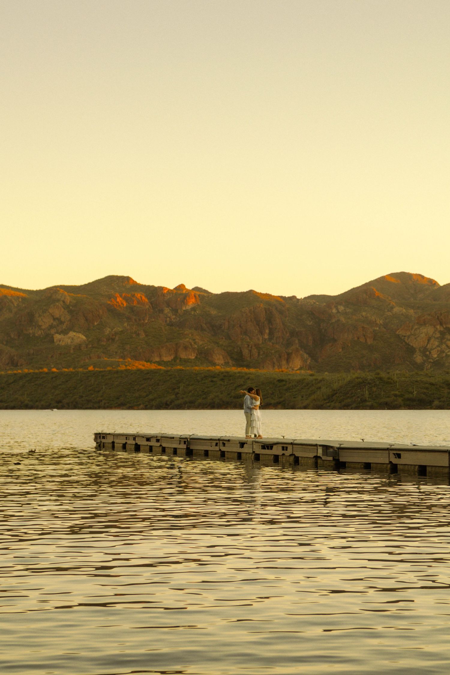 A couple stands together on a wooden dock at sunset with mountains silhouetted against the golden sky.