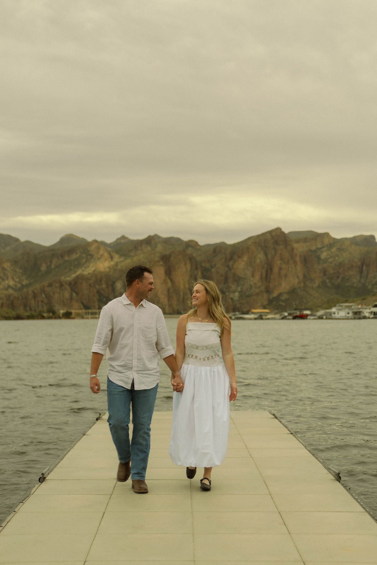 A couple in white outfits walks together on a dock overlooking a scenic mountain lake.