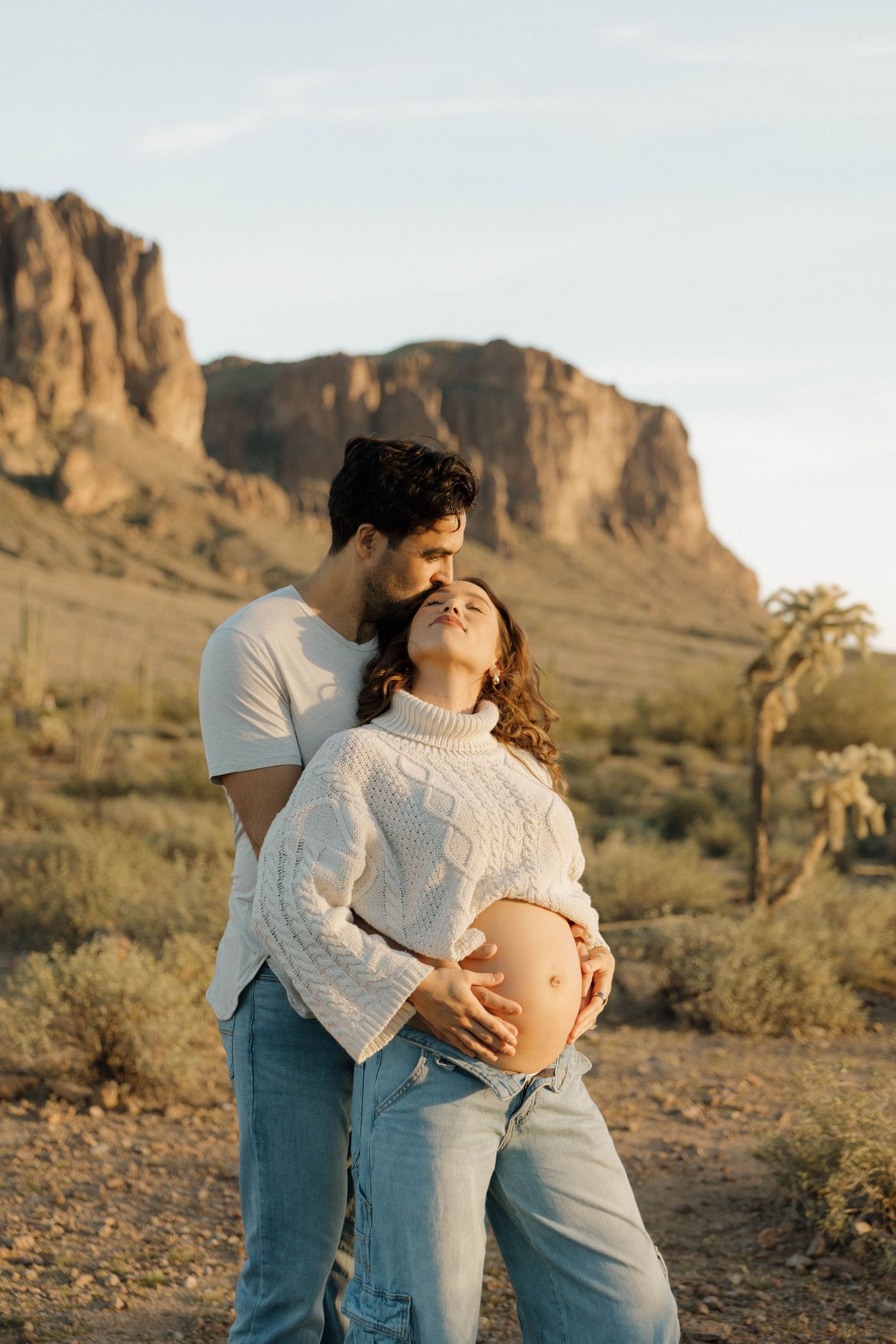 Maternity photoshoot in the desert with rugged mountain backdrop and couple embracing at sunset.