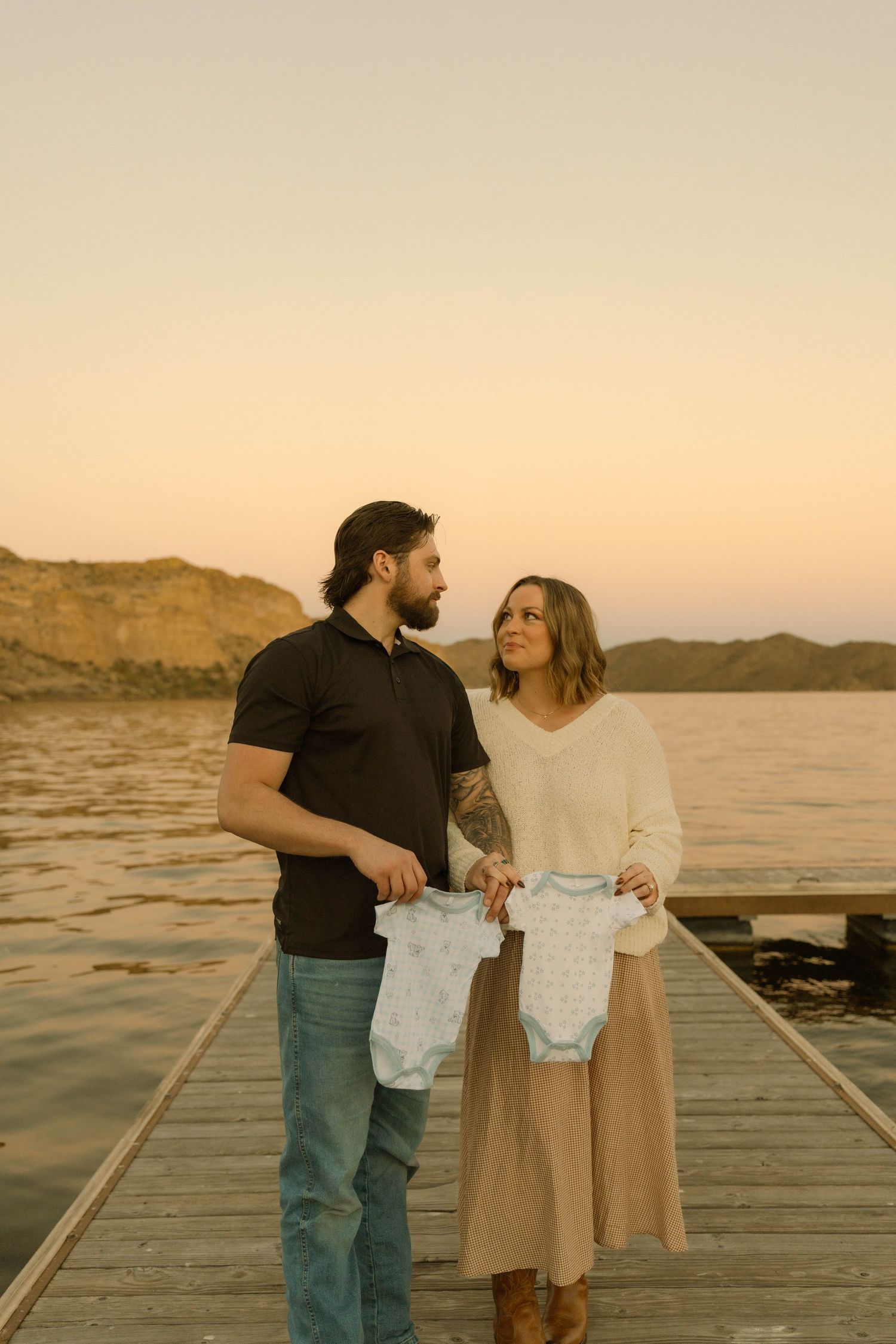 A couple stands on a dock at sunset holding baby clothes while looking at each other with joy and anticipation.