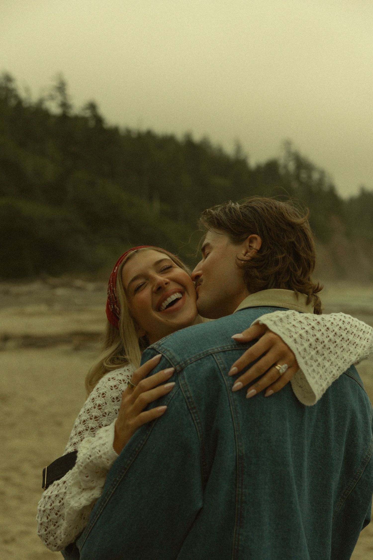 Couple sharing a romantic kiss on a misty beach, woman laughing joyfully in white sweater and red headband.