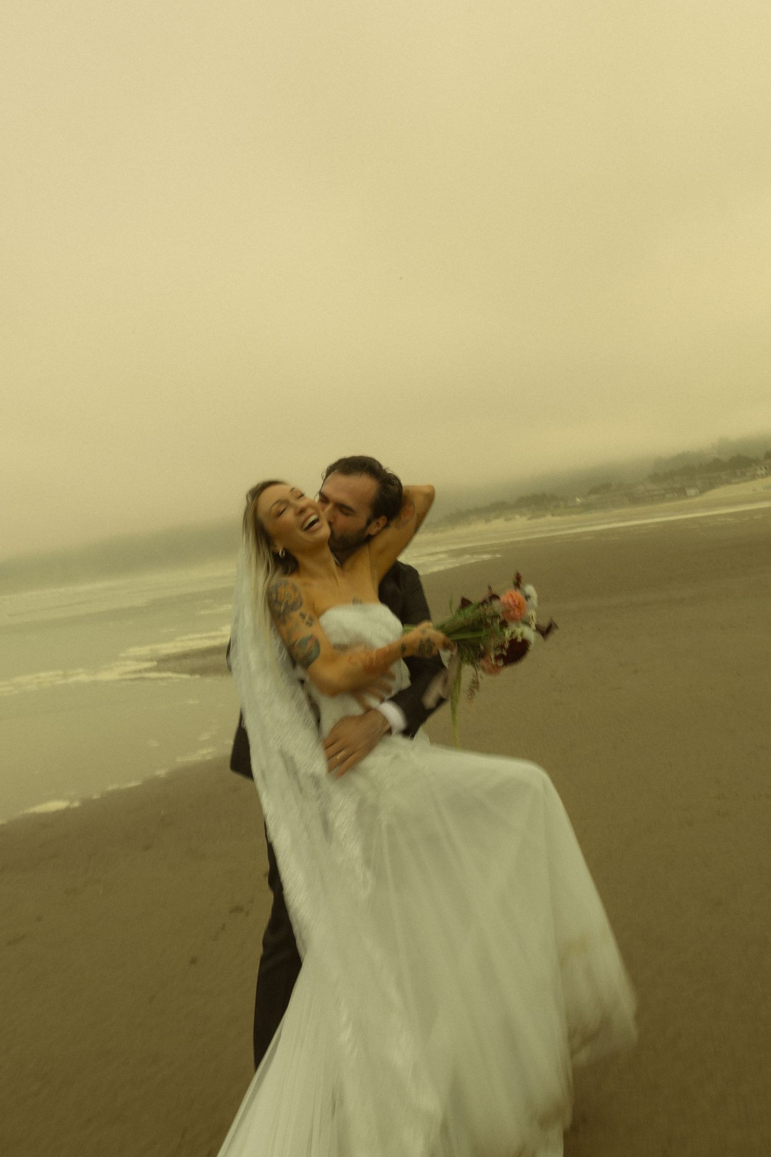 Joyful groom lifts laughing tattooed bride holding bouquet on a windy Oregon beach.