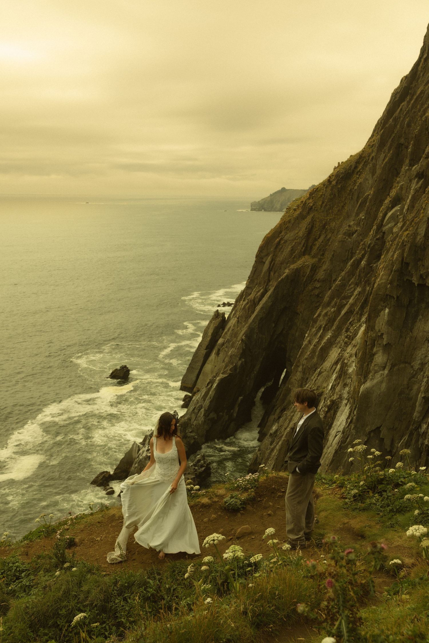 Couple in wedding attire standing on a dramatic coastal cliff overlooking the ocean with wildflowers.