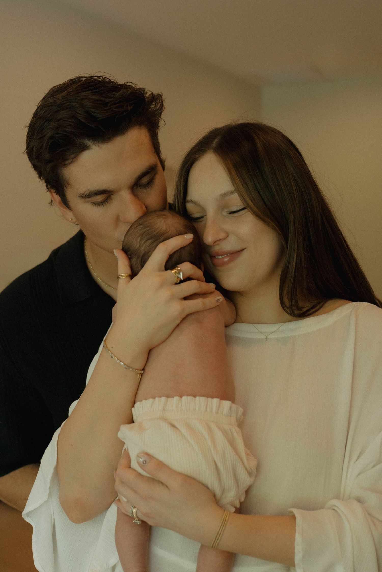 New parents tenderly hold their newborn baby, with father kissing the infant as mother smiles peacefully.