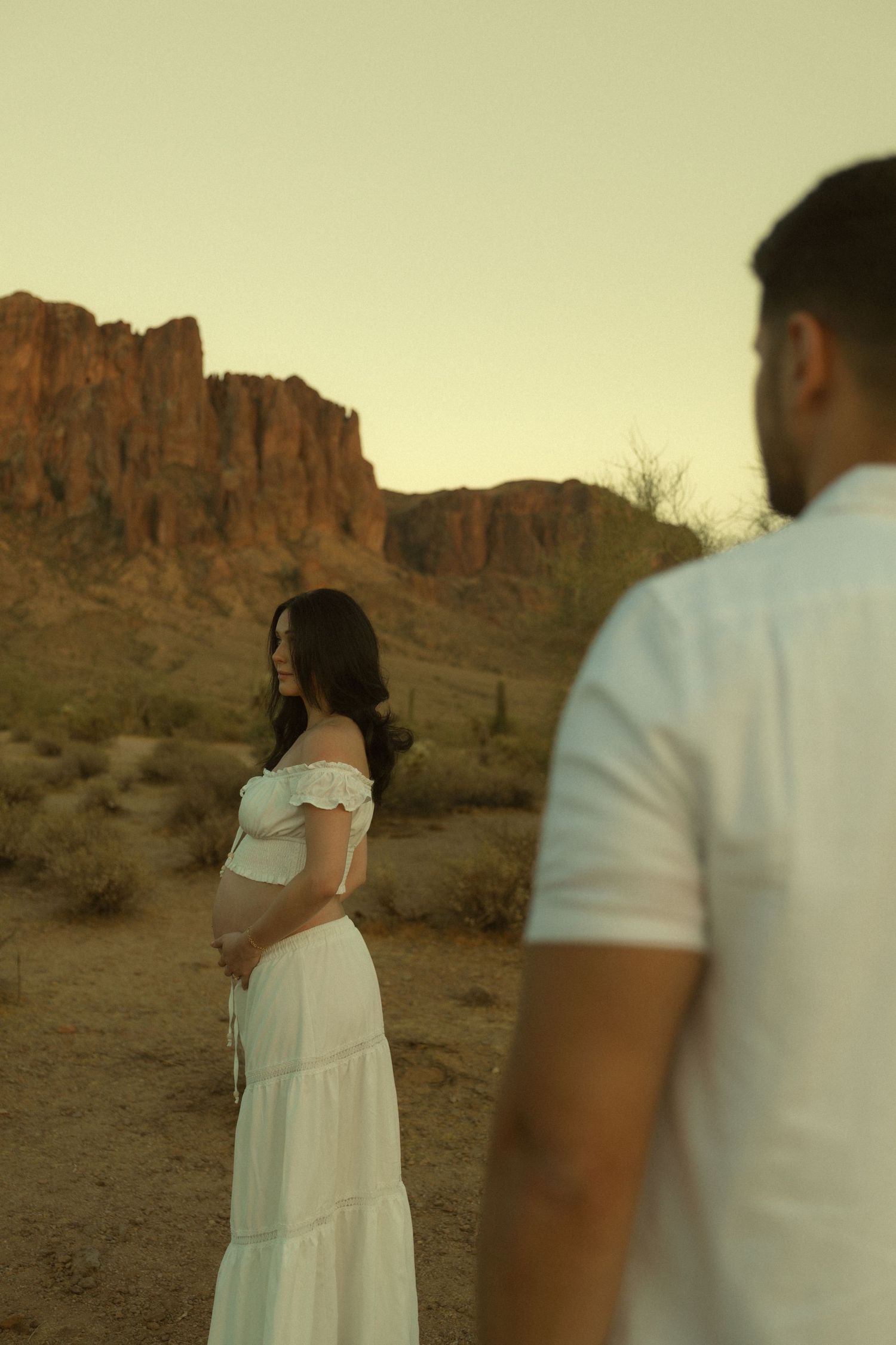 Pregnant woman in white off-shoulder dress holds baby bump in desert landscape with red rock formations at sunset.