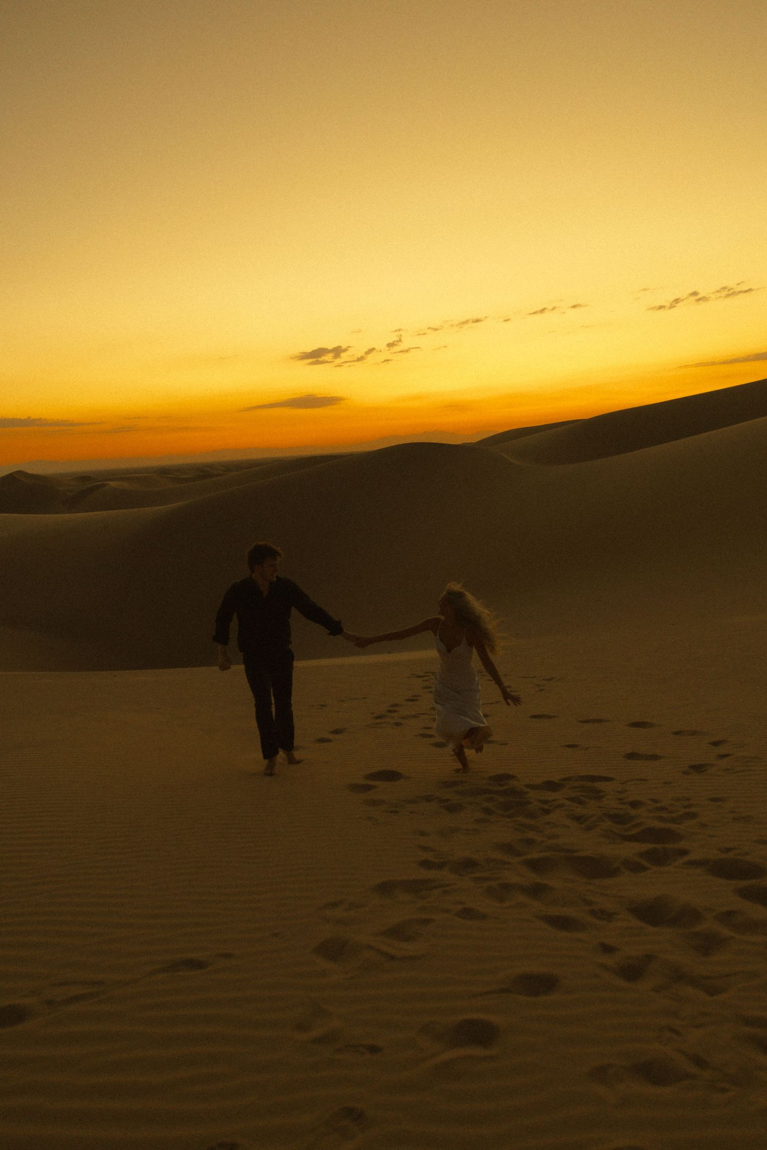 Silhouetted couple holding hands walking on sand dunes against a vivid orange desert sunset.