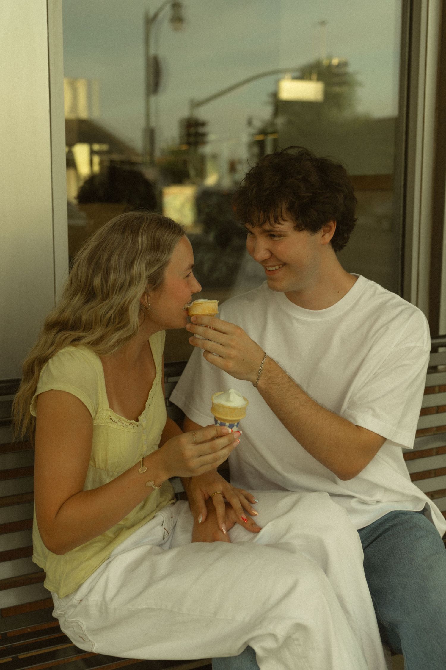 Young couple sharing ice cream cones on a bench, smiling at each other in a sweet romantic moment.