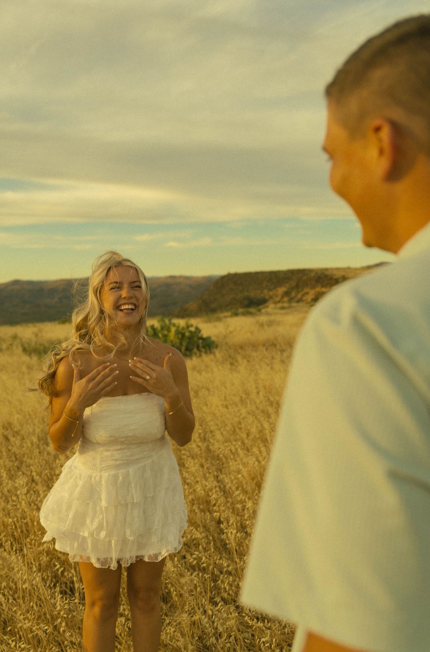A laughing woman in a white dress stands in a golden field, facing a man in a light blue shirt at sunset.