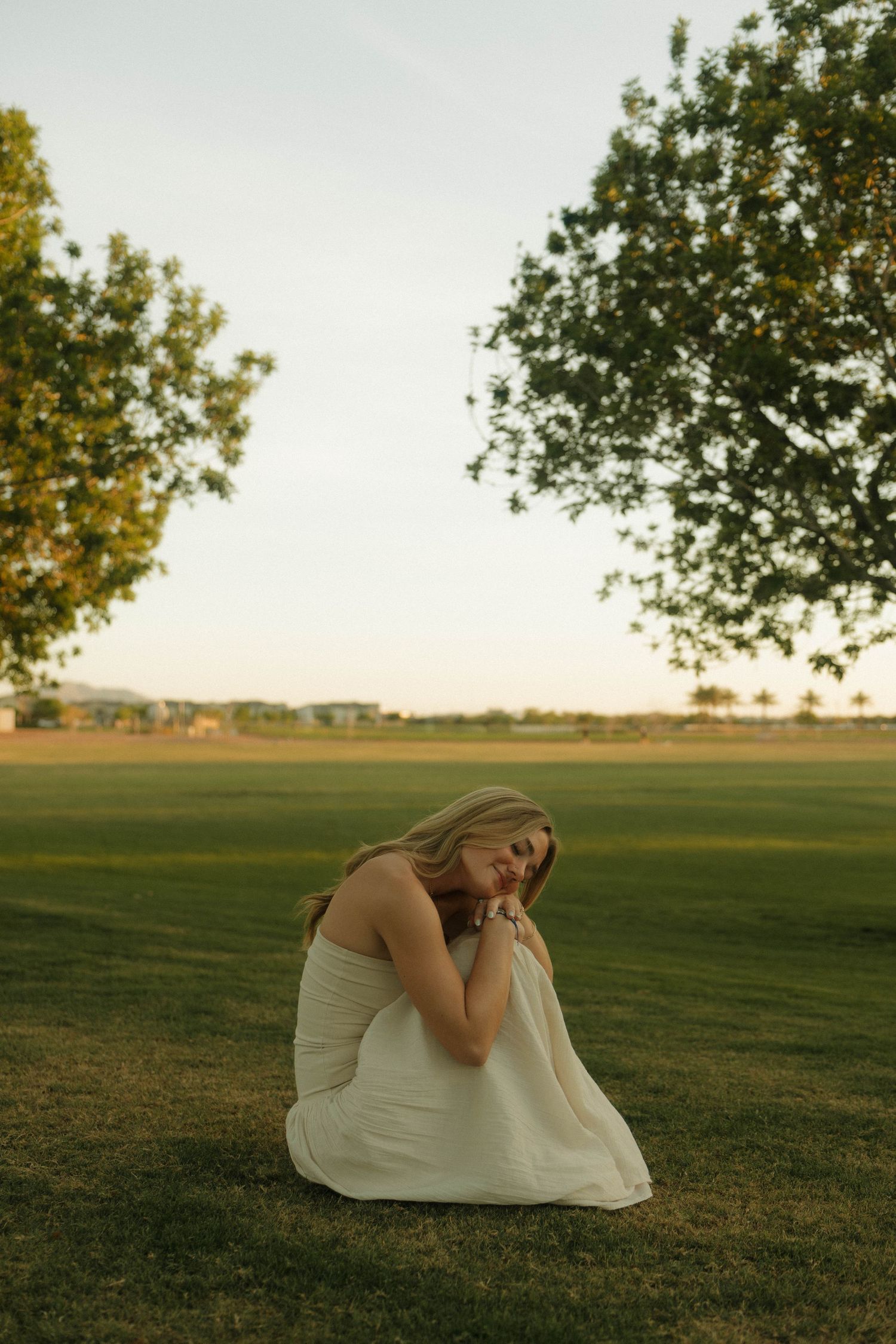 A woman in a white dress sits peacefully on green grass between two trees at golden hour sunset.
