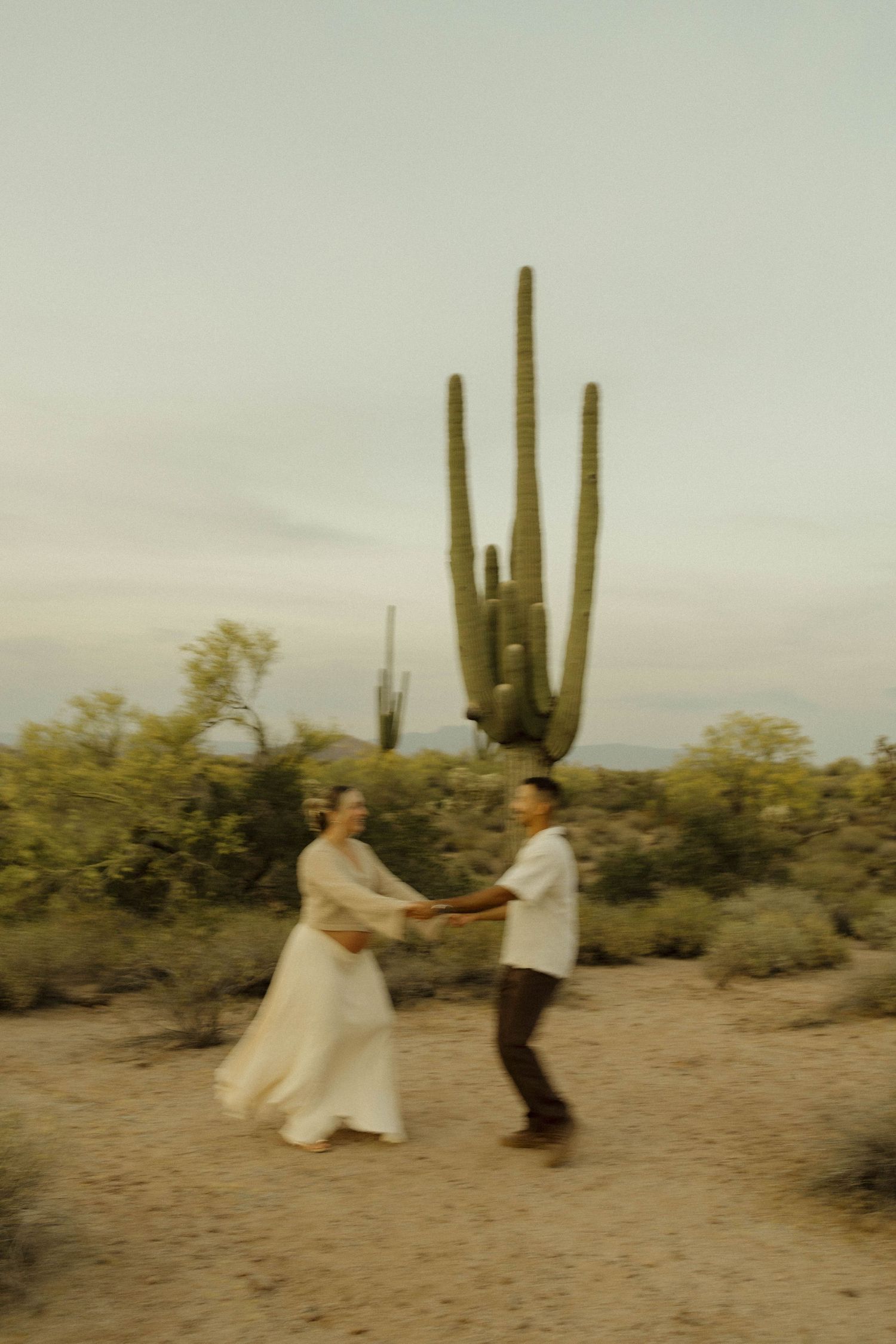 Couple dancing joyfully in Arizona desert with tall saguaro cactus backdrop during golden hour.