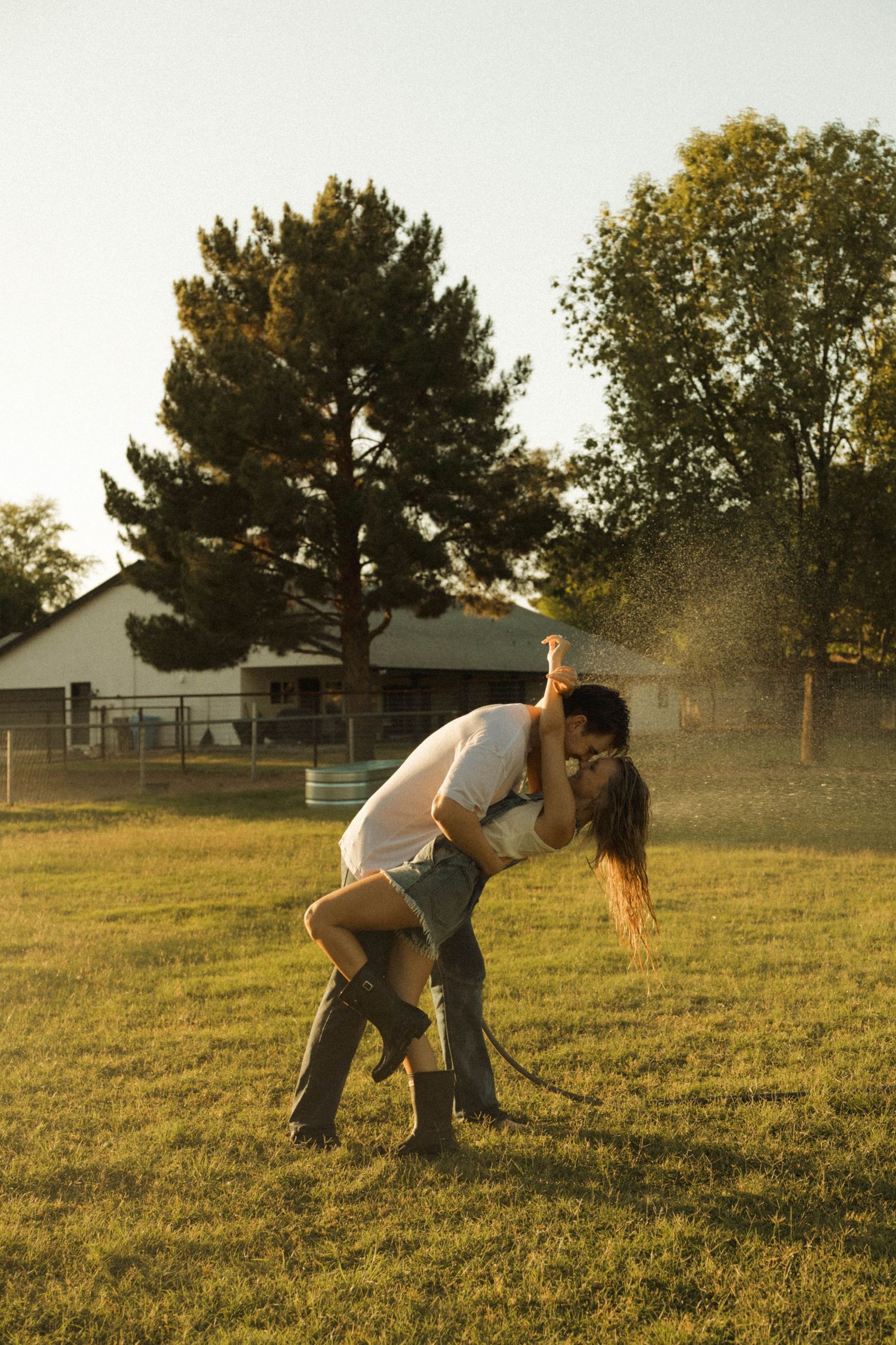 Couple kissing in a romantic dip pose on a farm lawn with water hose spray at golden hour.