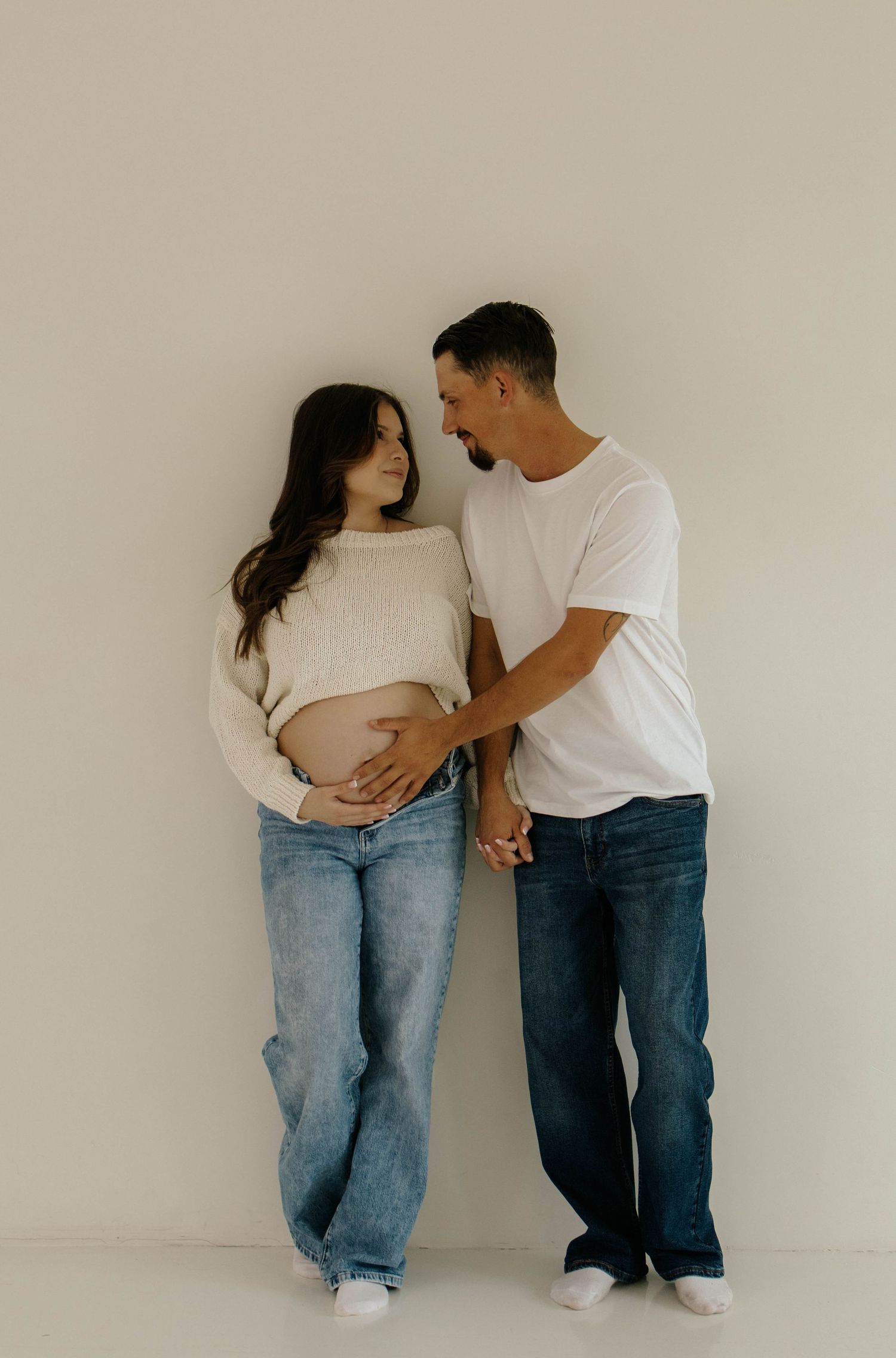 A couple in matching white shirts and jeans share tender moments during a minimalist maternity photoshoot against a neutral wall.