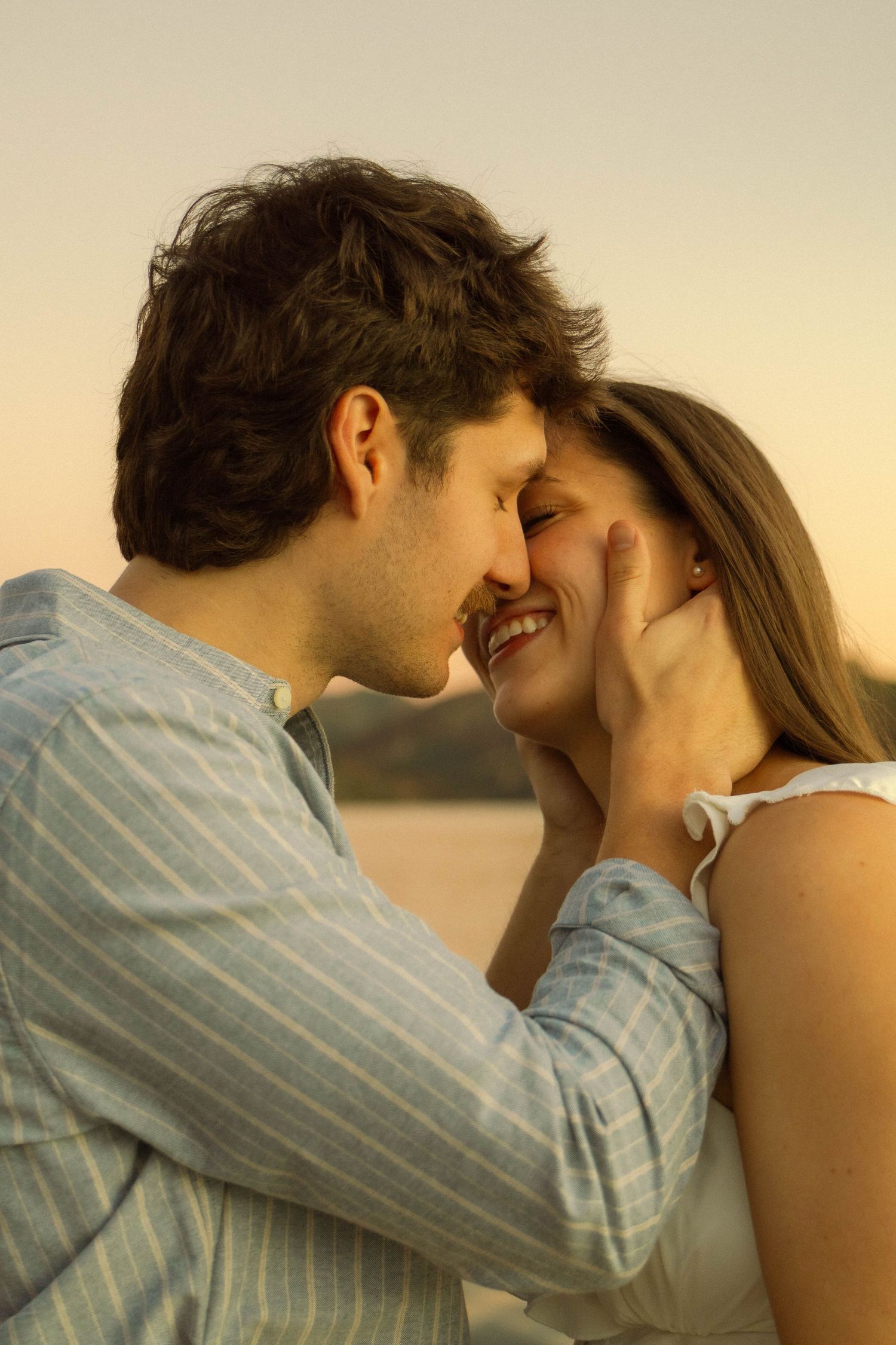 Tender moment between couple in casual attire against sunset backdrop near water.