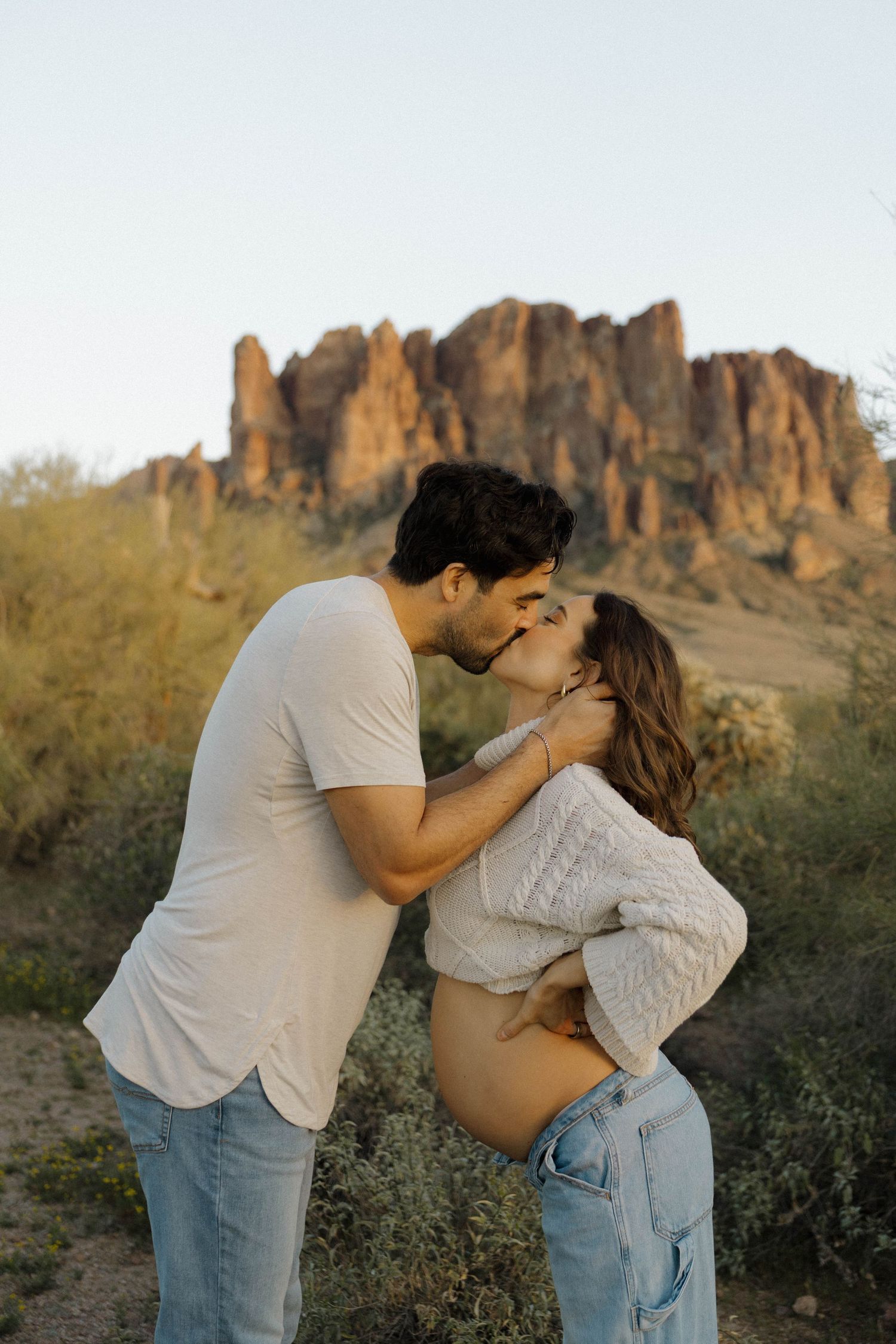 Couple embraces and kisses in front of scenic desert mountain landscape during golden hour.
