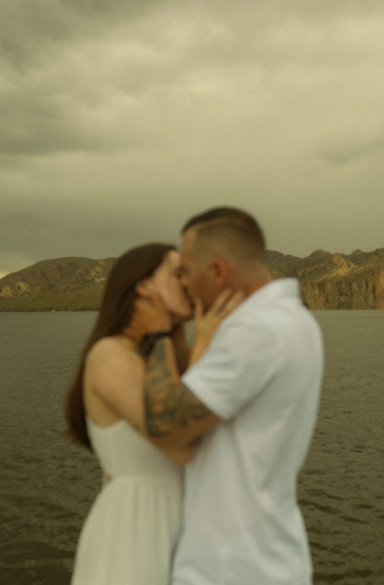 A romantic couple shares an intimate moment by a scenic lake with mountains in the background at sunset.