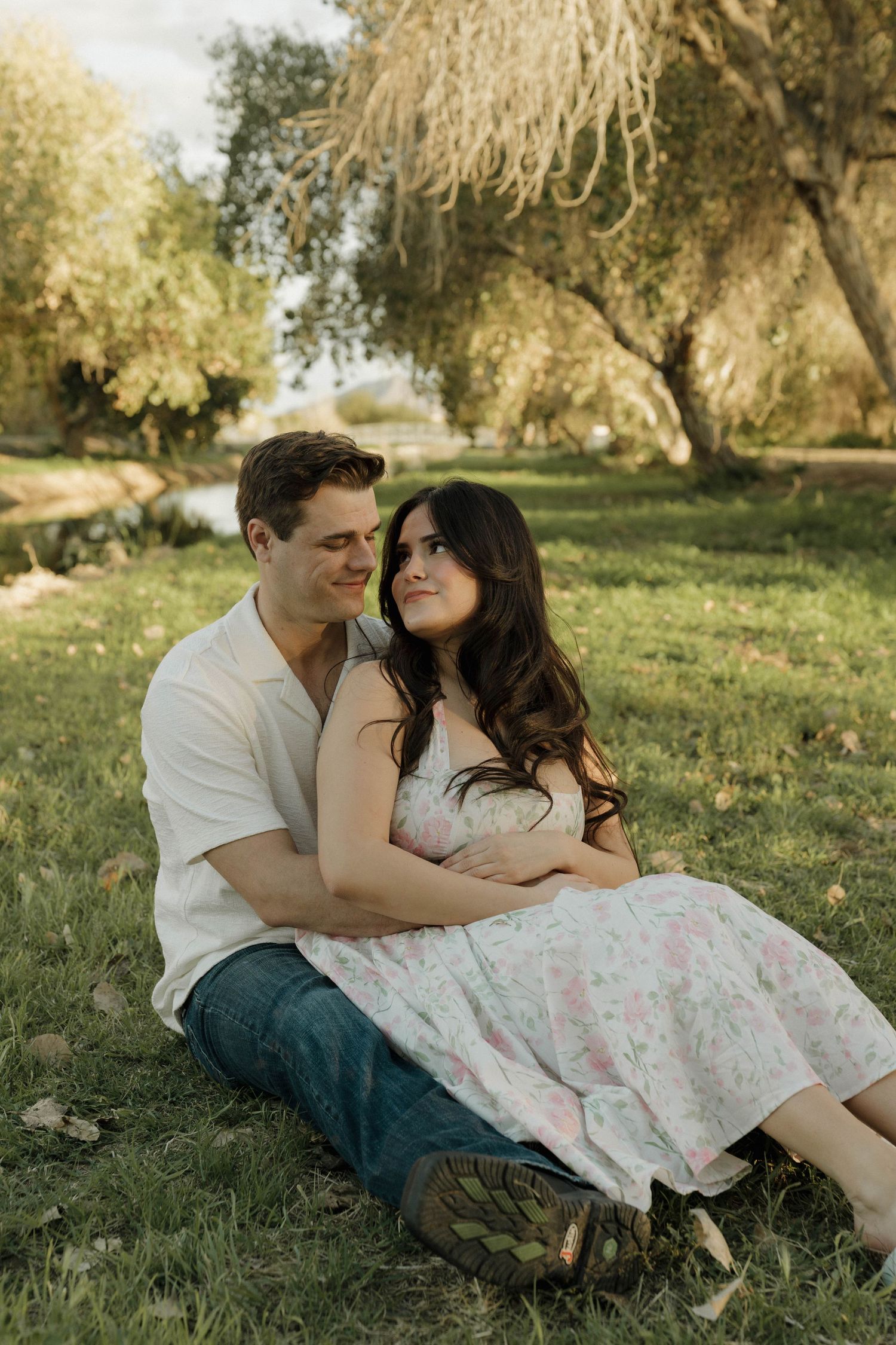 A romantic couple lounges on grassy hillside during golden hour beneath willow trees.