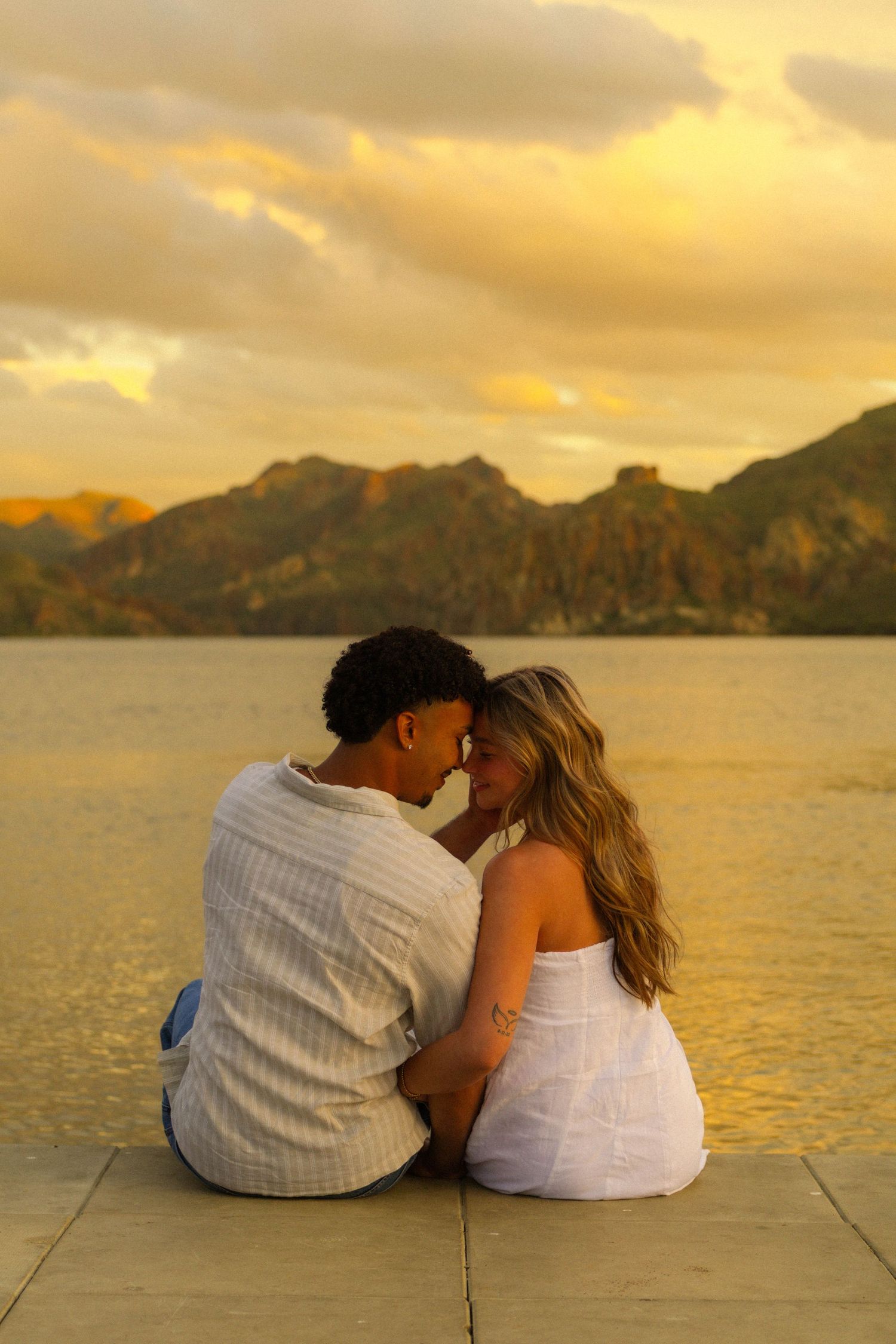 A couple sits together watching a golden sunset over a mountainous bay during a romantic evening.