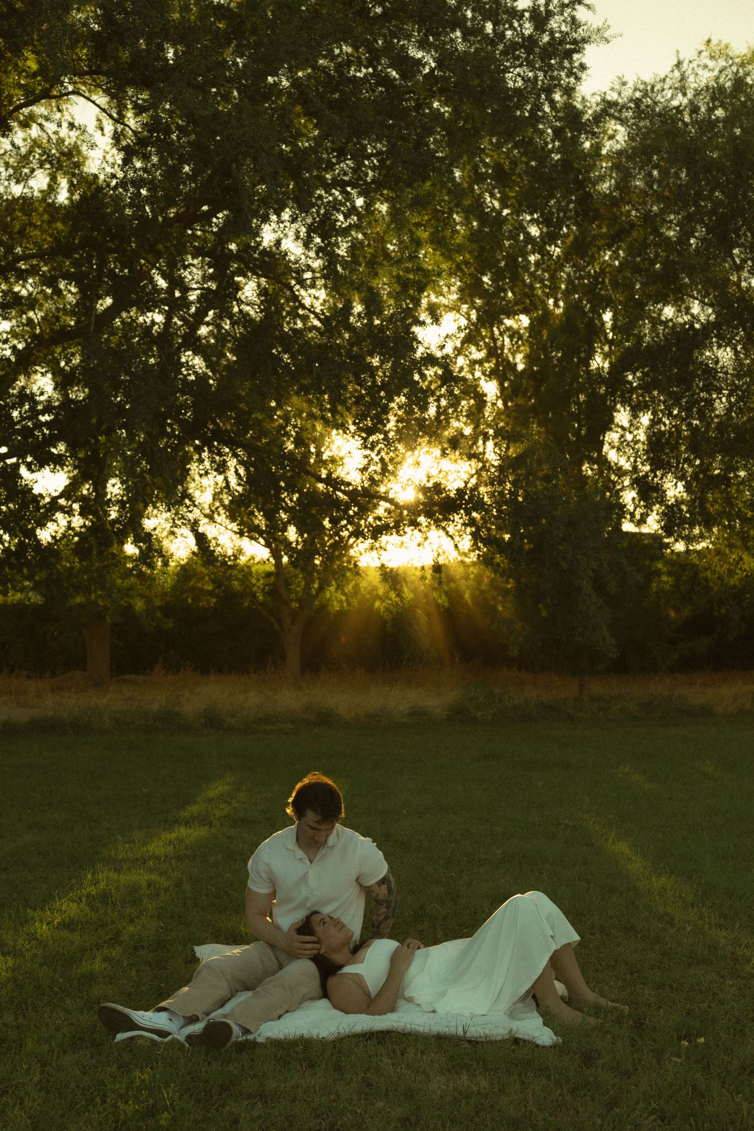 Couple relaxing on a blanket in a sunlit green field at golden hour with trees glowing behind them.