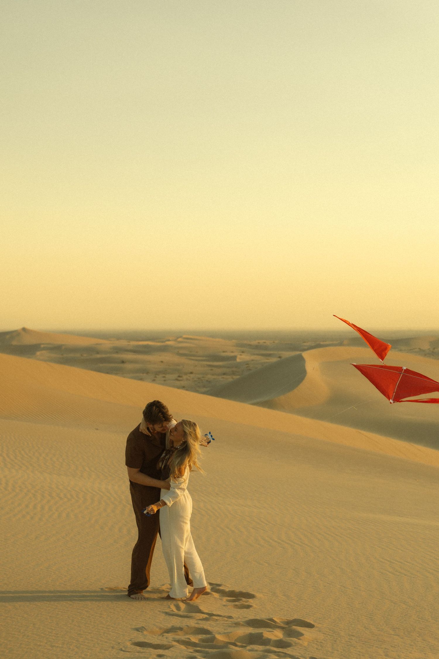 Couple embracing romantically on golden sand dunes at sunset with a red kite flying in the desert sky.