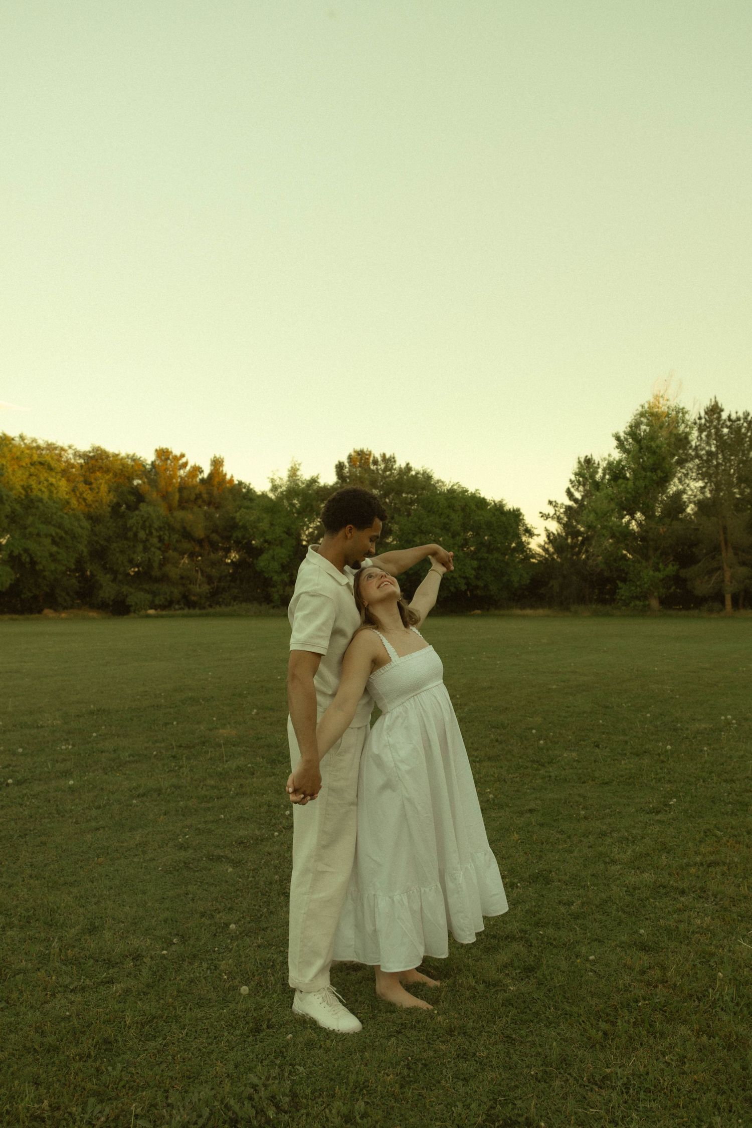 A couple in white outfits dancing romantically in a lush green park at golden hour sunset.