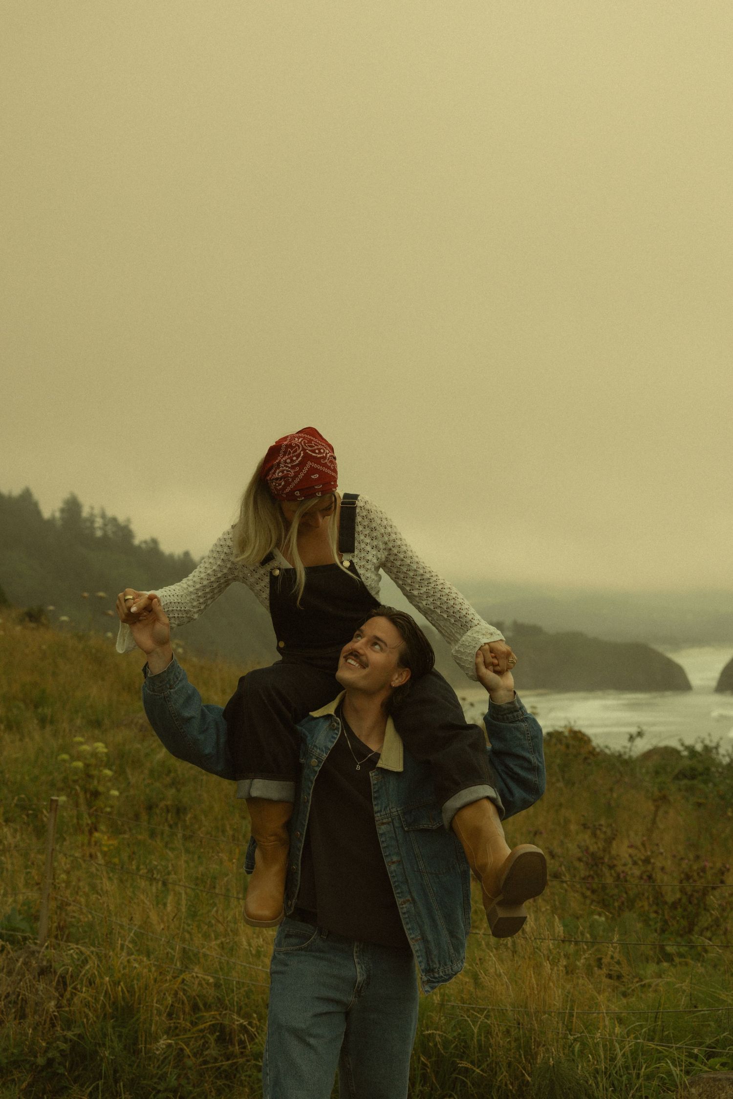 A smiling woman carries another person on her shoulders in a misty coastal meadow, both joyful outdoors.