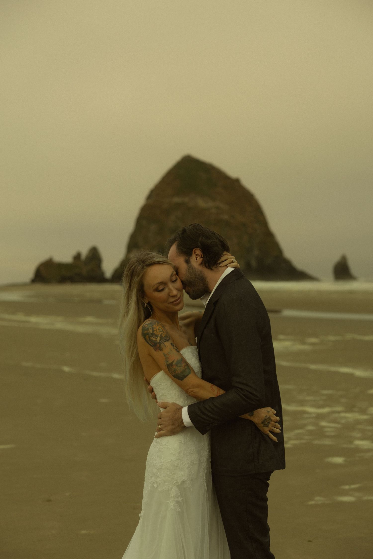 Bride and groom share a romantic kiss on the beach with Haystack Rock in the background at Cannon Beach, Oregon.