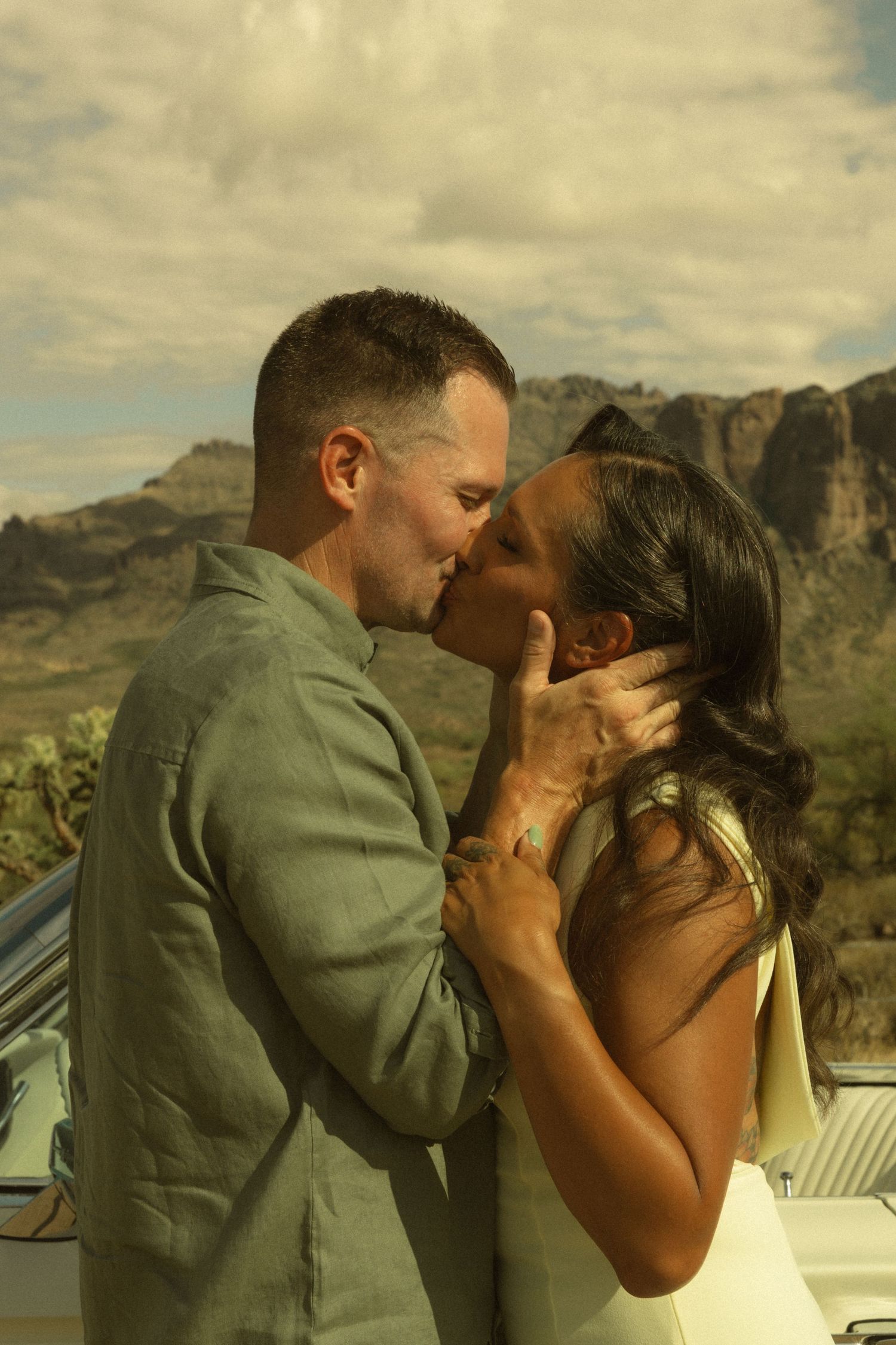 Couple sharing a romantic kiss in desert landscape with mountains, wearing wedding attire near vintage car.