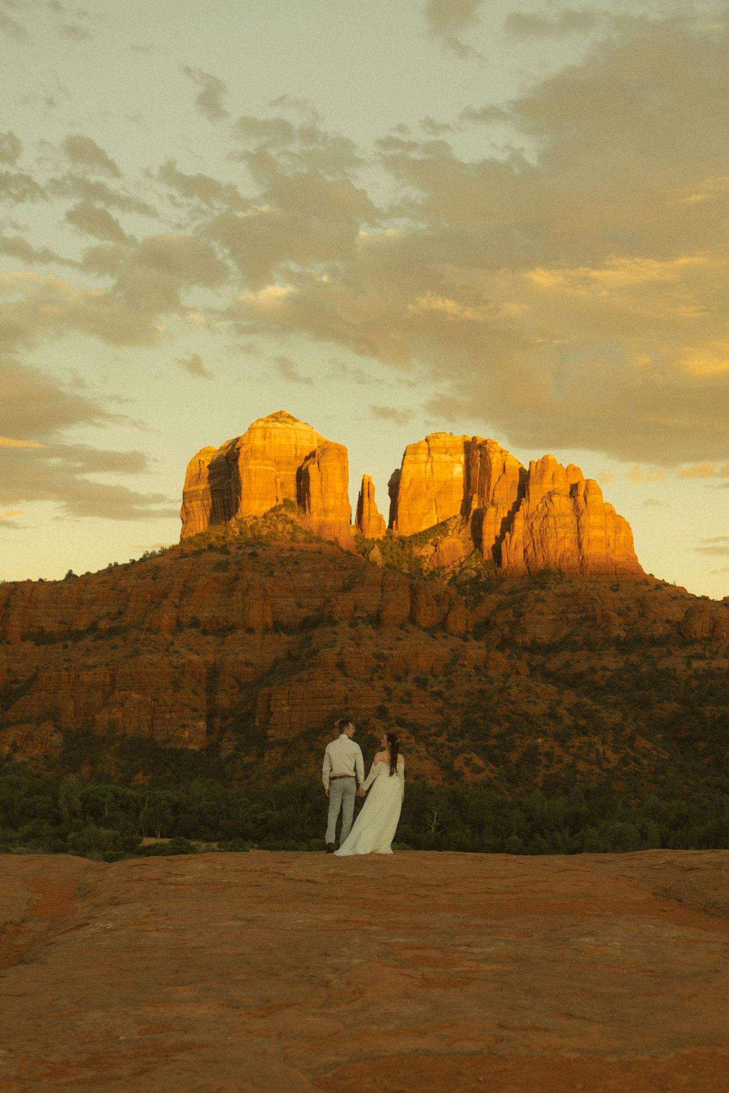 Couple in wedding attire standing before Cathedral Rock at golden sunset in Sedona, Arizona red rock landscape.