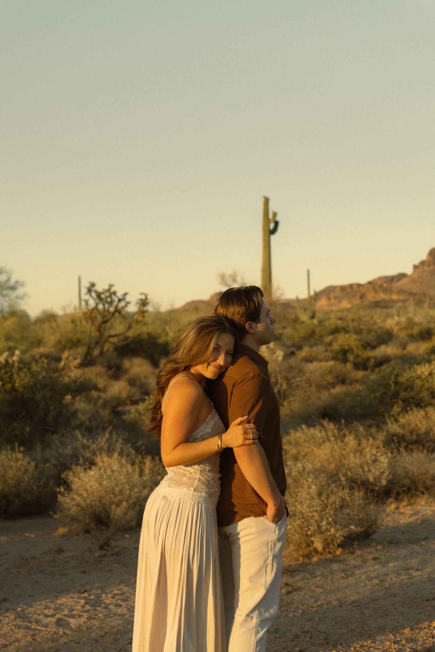 Couple holding each other close in the Arizona desert at sunset with saguaro cacti and mountains behind them.