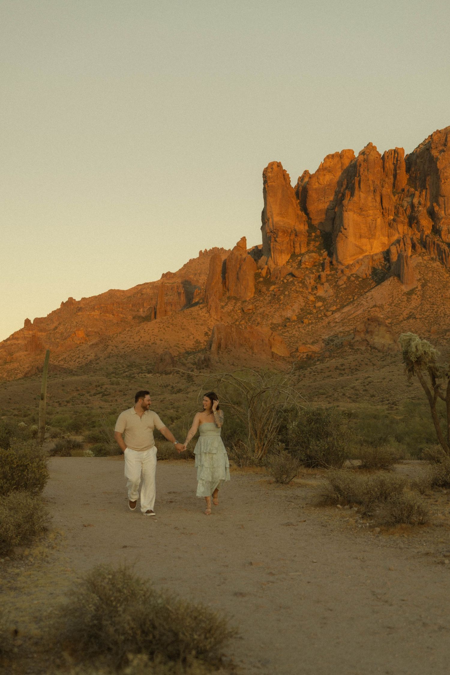 Couple holding hands walking in Arizona desert at golden hour with Superstition Mountains glowing red behind them.