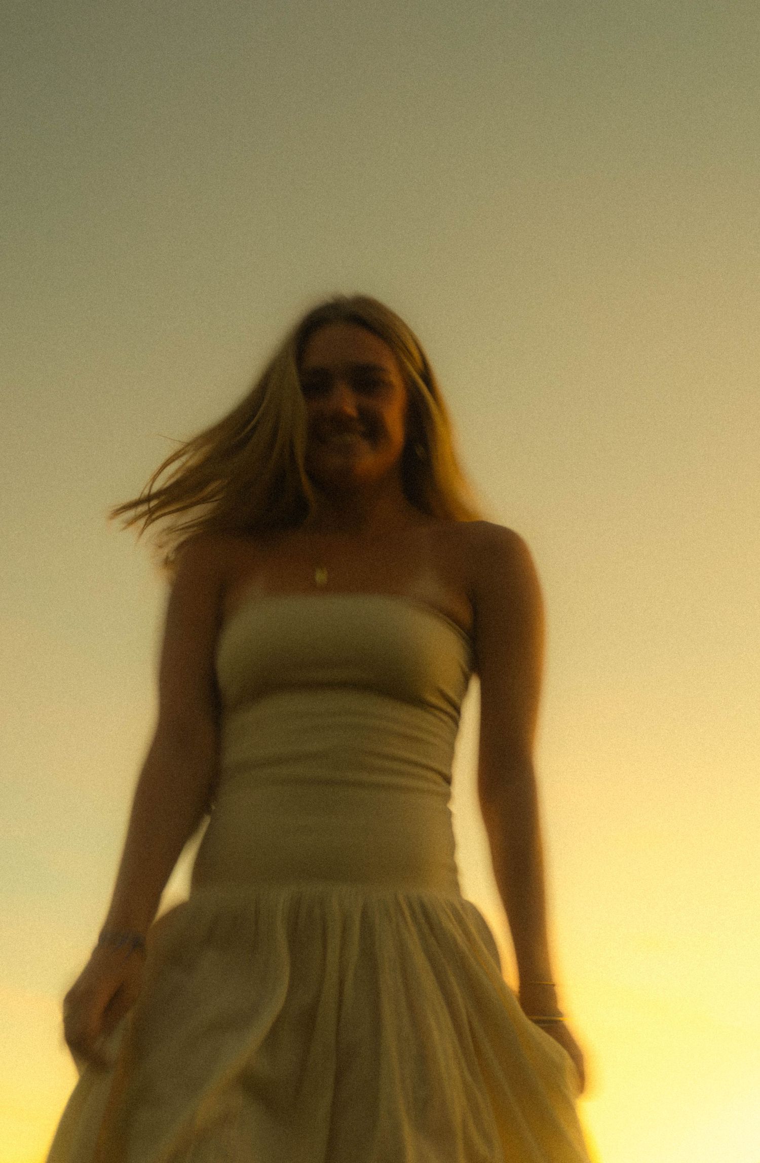 Young woman in white strapless dress smiling at sunset with golden sky background and hair blowing in the wind.