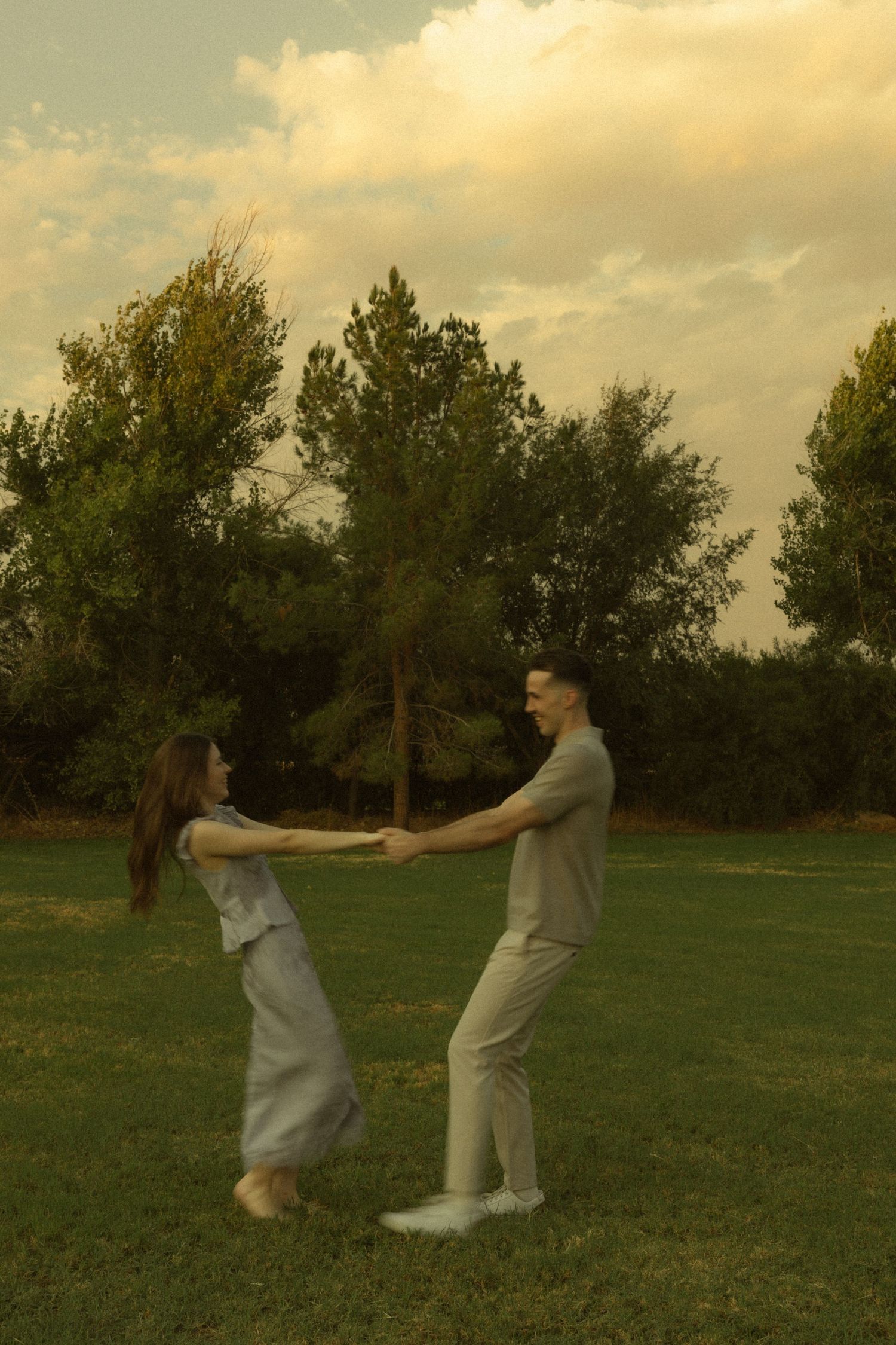 A couple joyfully spinning together on a green lawn at sunset with lush trees in the background.