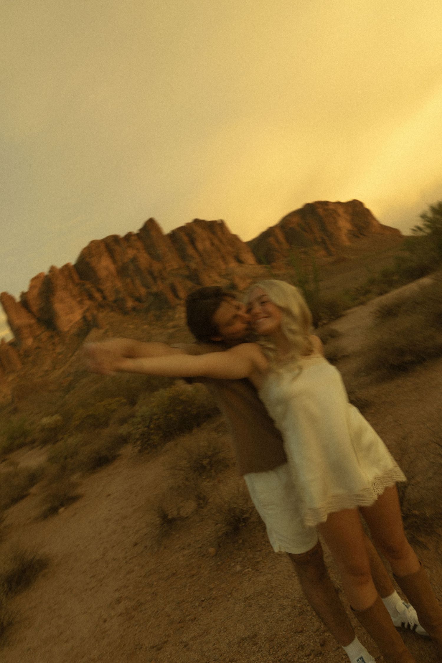 Two friends embrace and spin joyfully in the desert at golden sunset with rocky mountains in the background.