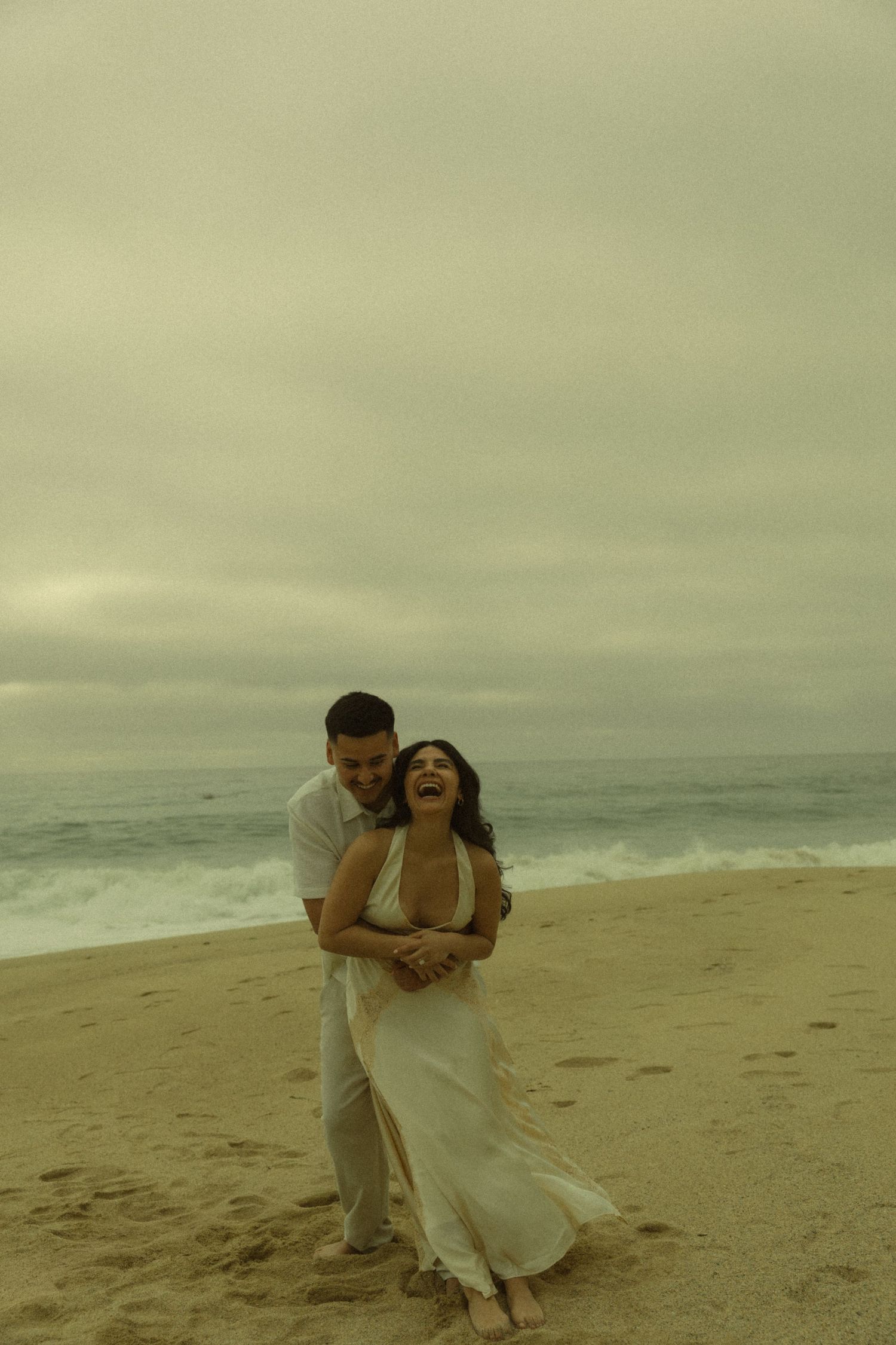 Couple laughing and embracing on a sandy beach with overcast skies and ocean waves behind them.