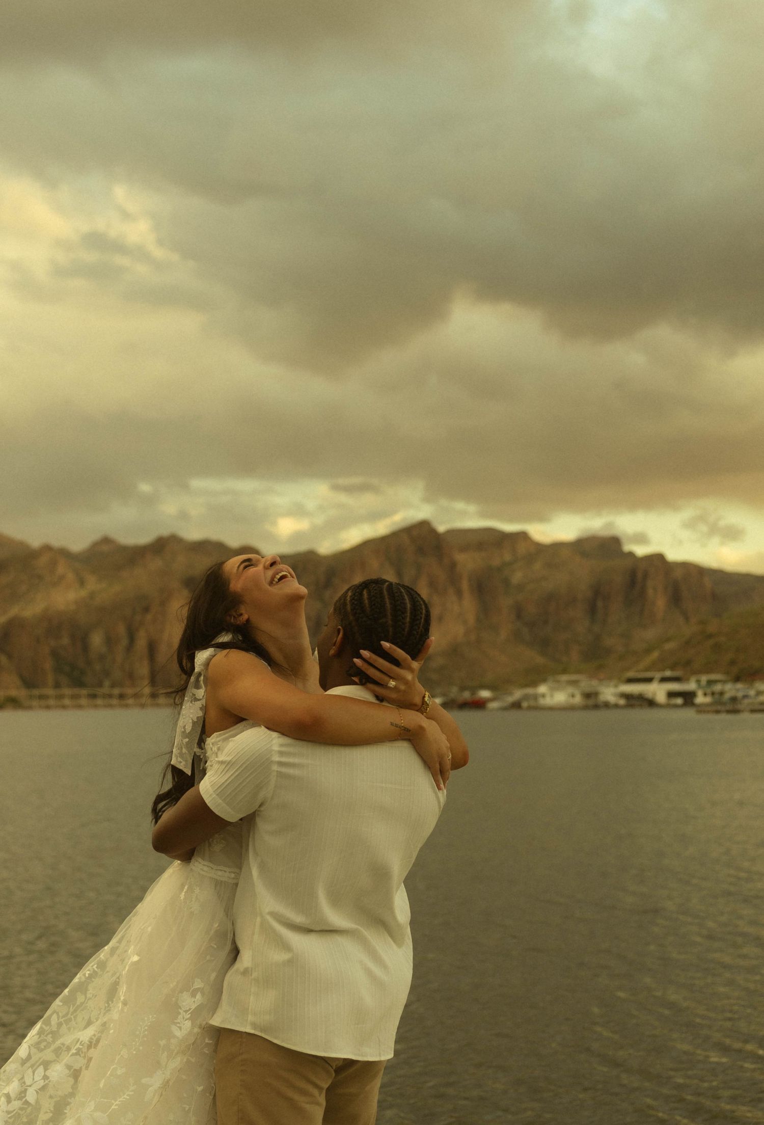 Couple in white embracing and laughing by a lake with dramatic mountain backdrop at golden sunset.
