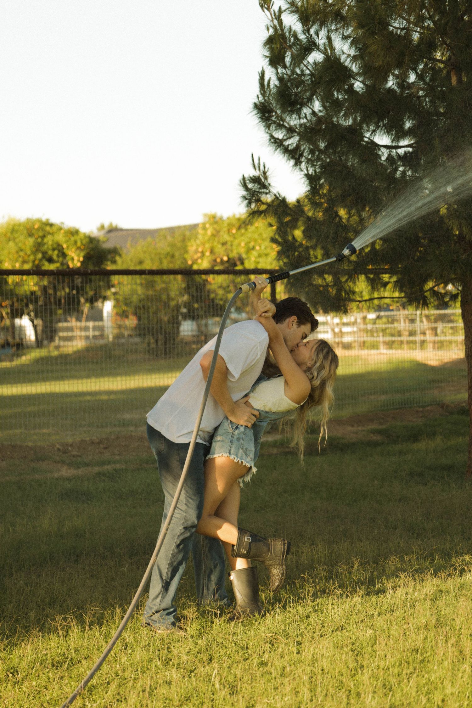A couple sharing a romantic kiss in a sunny backyard while holding a garden hose sprinkler.