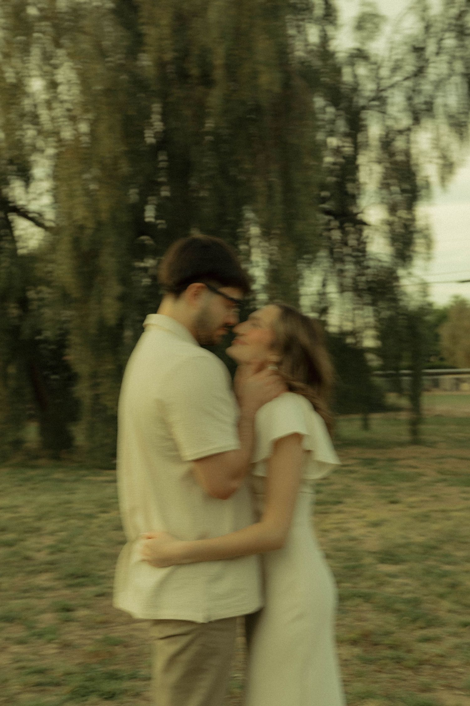 Couple embracing and kissing outdoors in a park, dressed in neutral tones, surrounded by willow trees in soft focus.