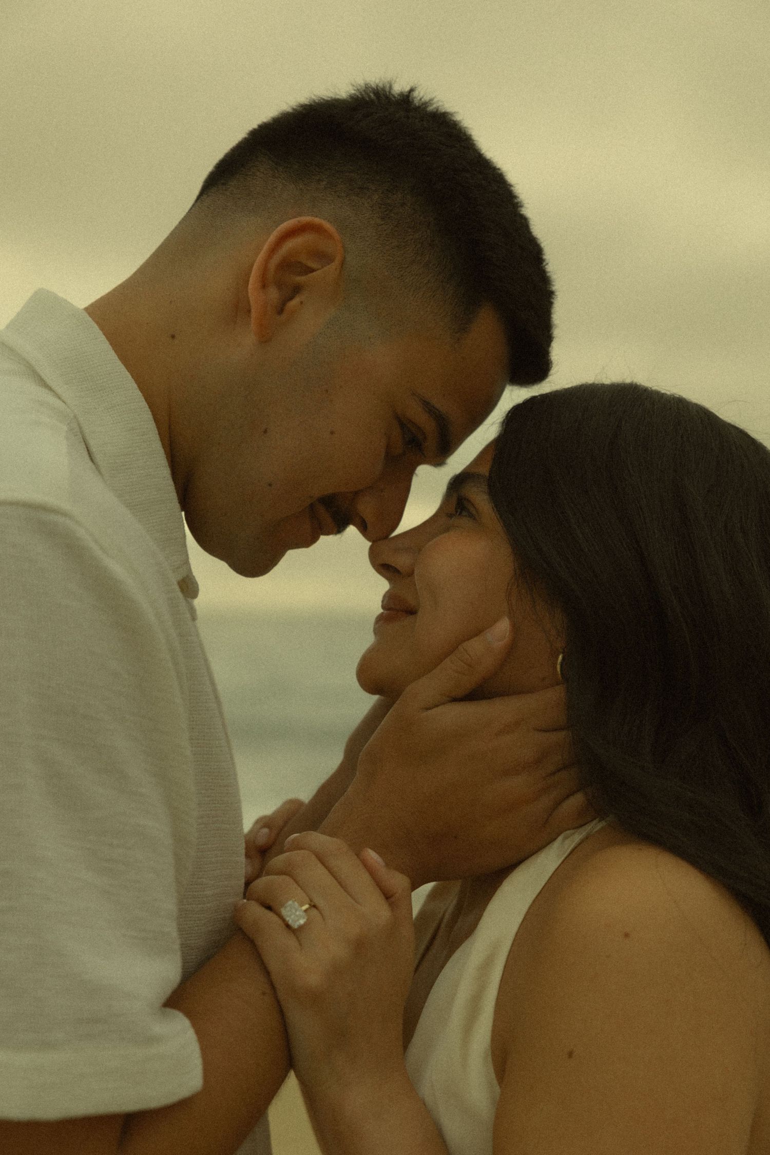 Couple sharing a romantic beach moment, foreheads touching, as man gently holds womans face, engagement ring visible.