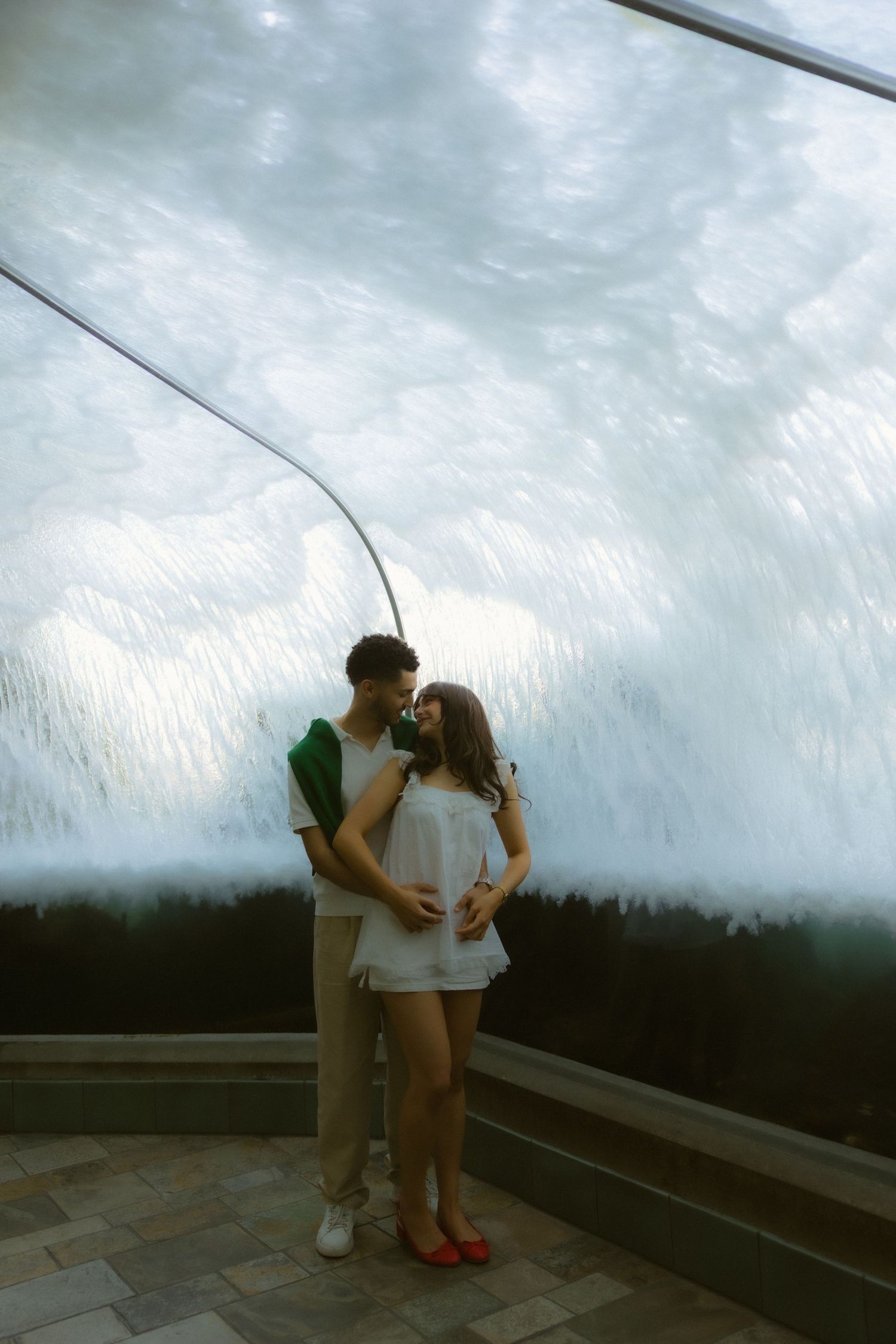 Romantic couple embracing in front of a large waterfall feature, woman in white dress and man in casual attire.