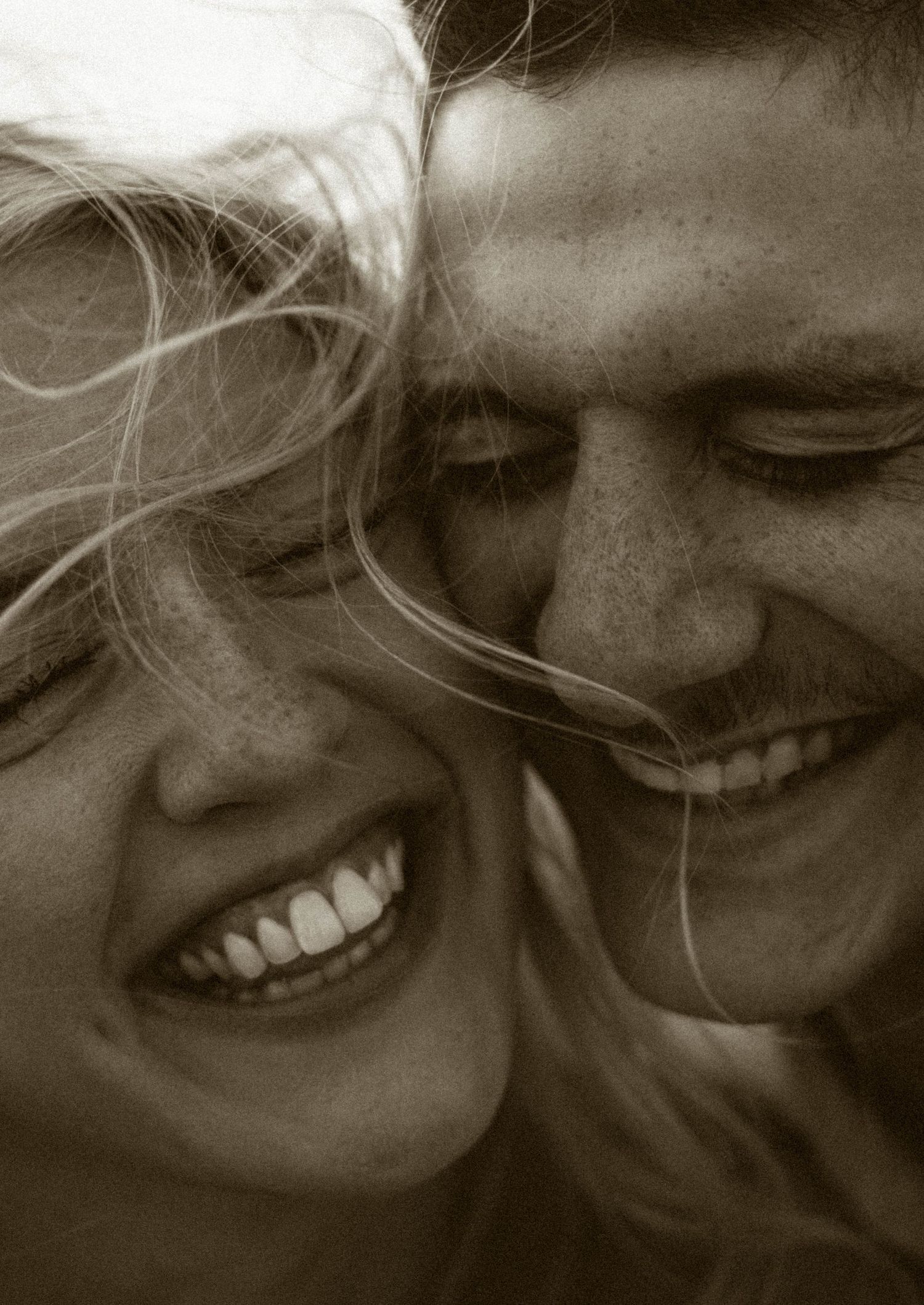 A joyful couple laughing together in a close sepia-toned portrait, woman with blonde hair and man with freckles.