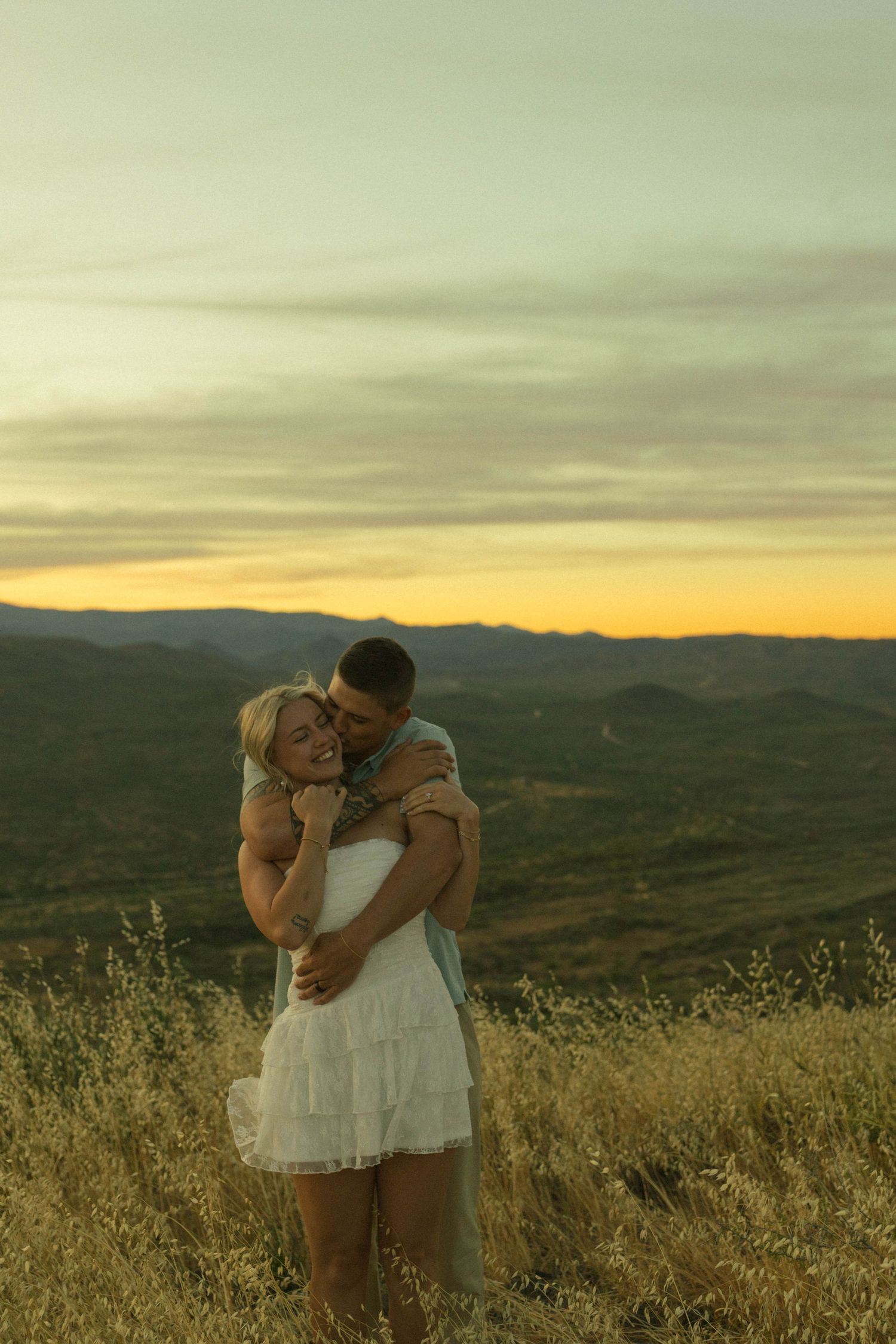 Romantic couple embracing at golden sunset with scenic mountain landscape behind.