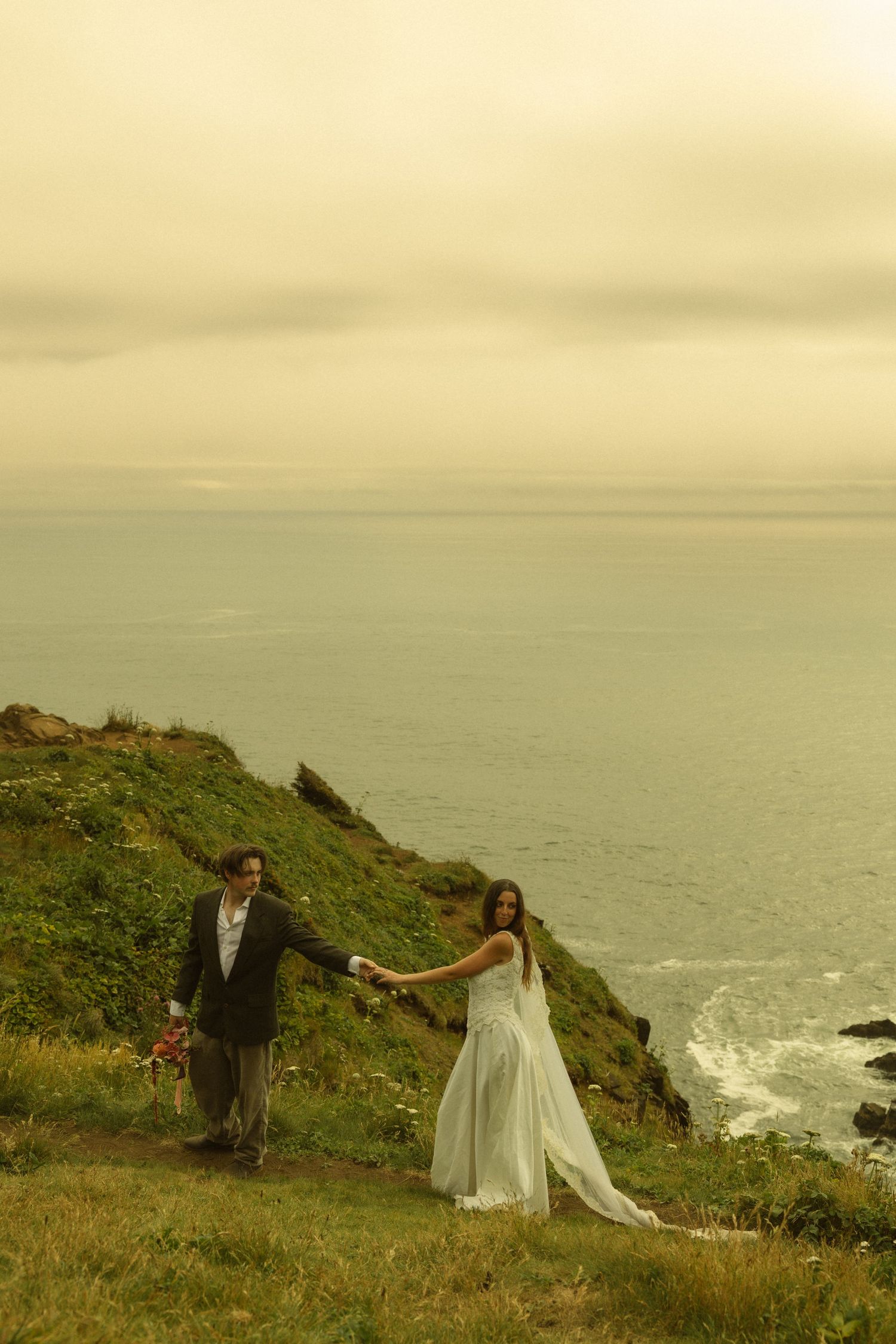 Wedding couple holds hands while walking along a lush green coastal cliff with ocean waves below.