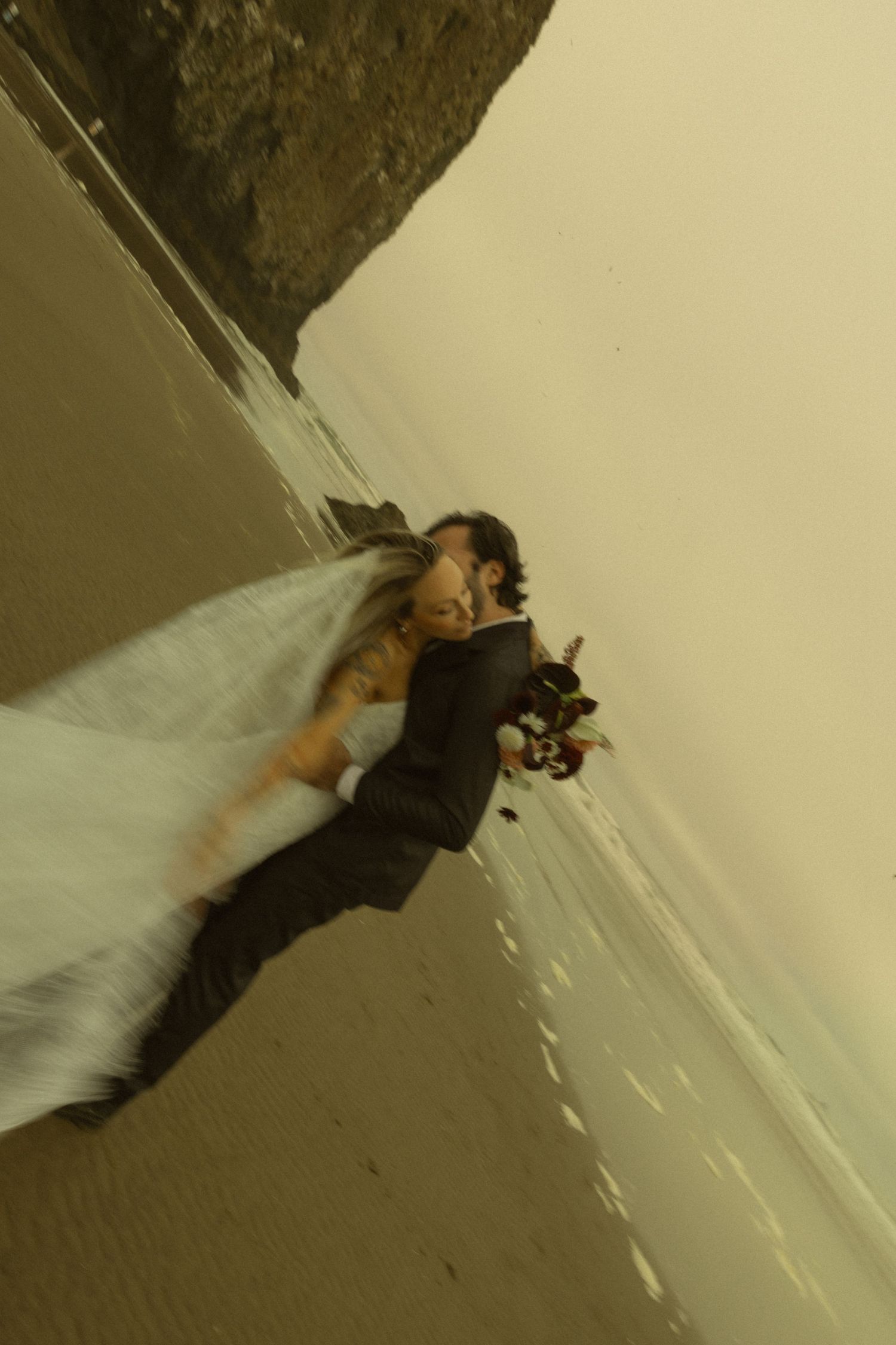 Bride and groom kissing on a sandy beach with ocean waves, wedding dress veil flowing in the wind.