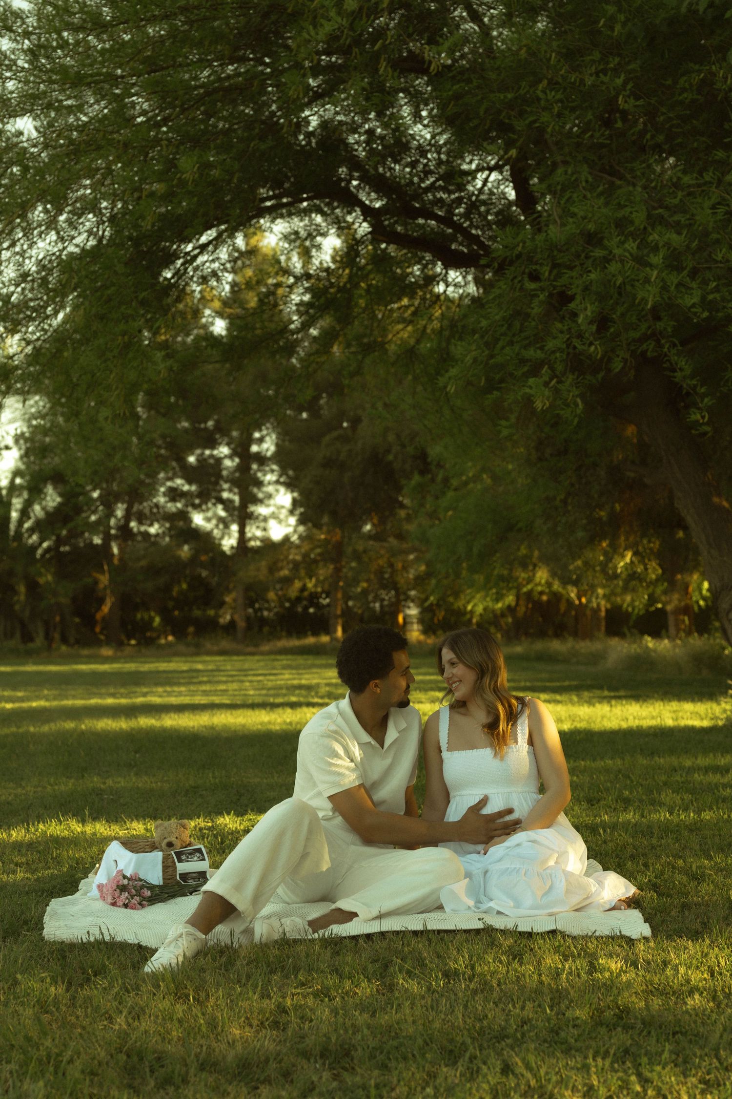 Couple sitting on blanket in park, man touching pregnant woman's belly, surrounded by lush green trees in golden light.
