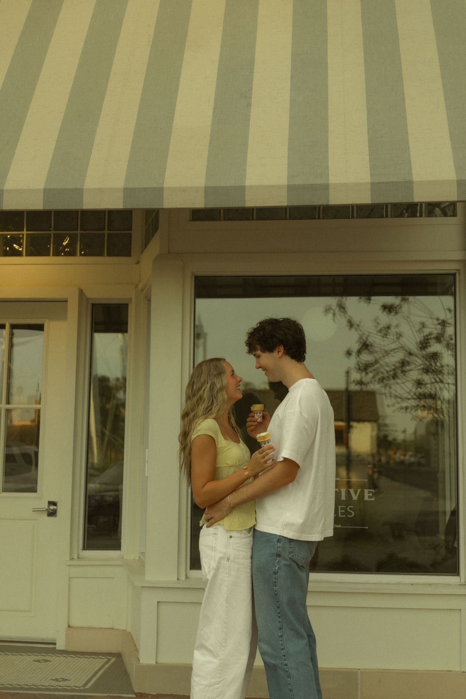Couple sharing an ice cream cone outside a striped awning storefront on a sunny day.